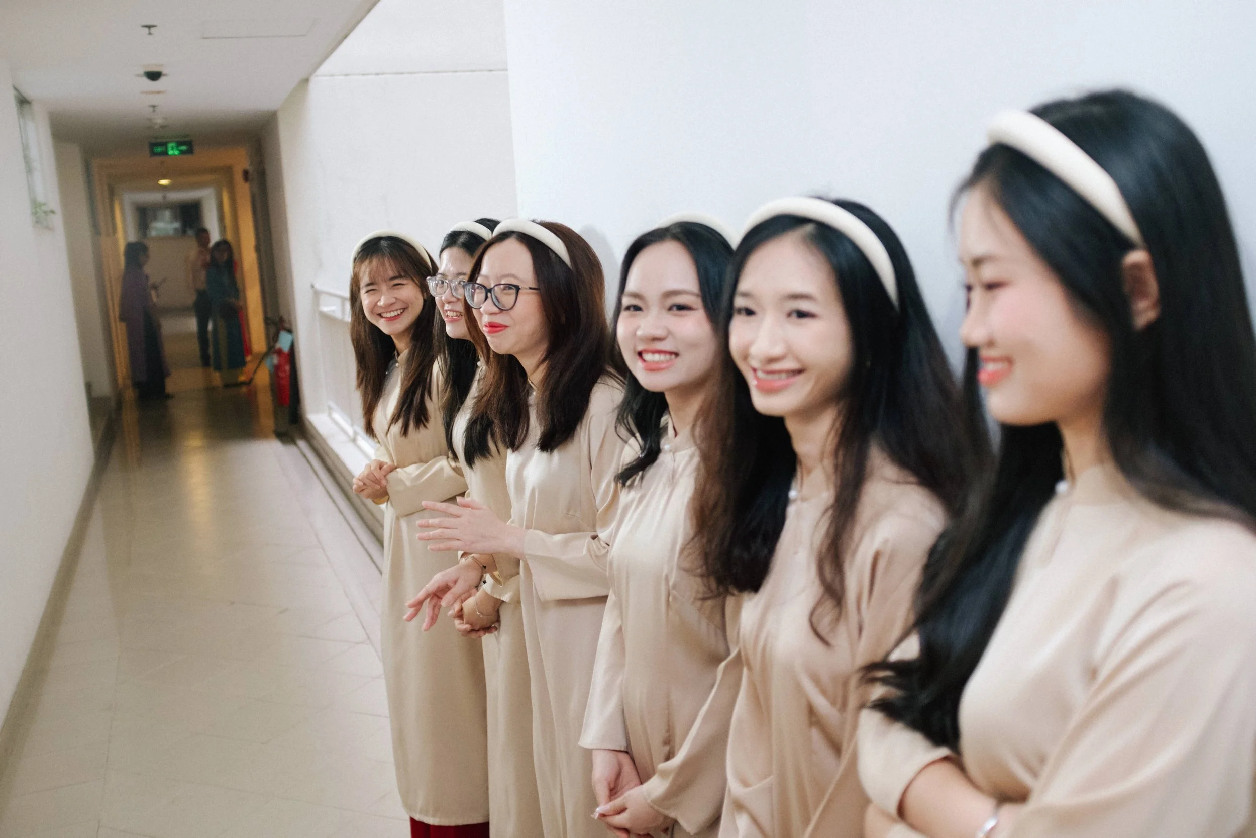 Seven women standing in a line along a corridor, smiling and wearing beige dresses with white headbands.