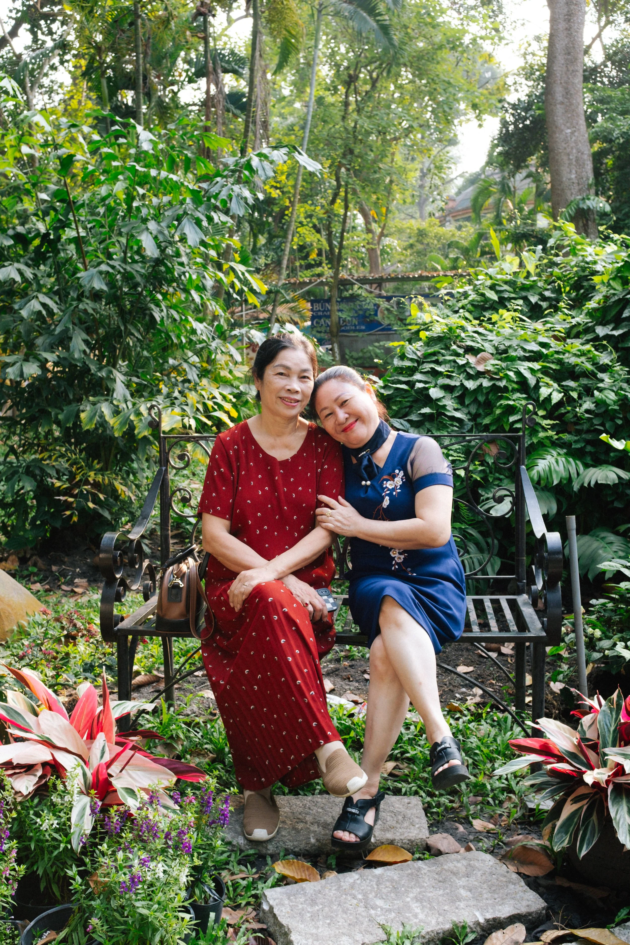 Two women sit on a black metal garden bench surrounded by lush greenery and colorful plants, smiling and leaning on each other in a tropical garden.