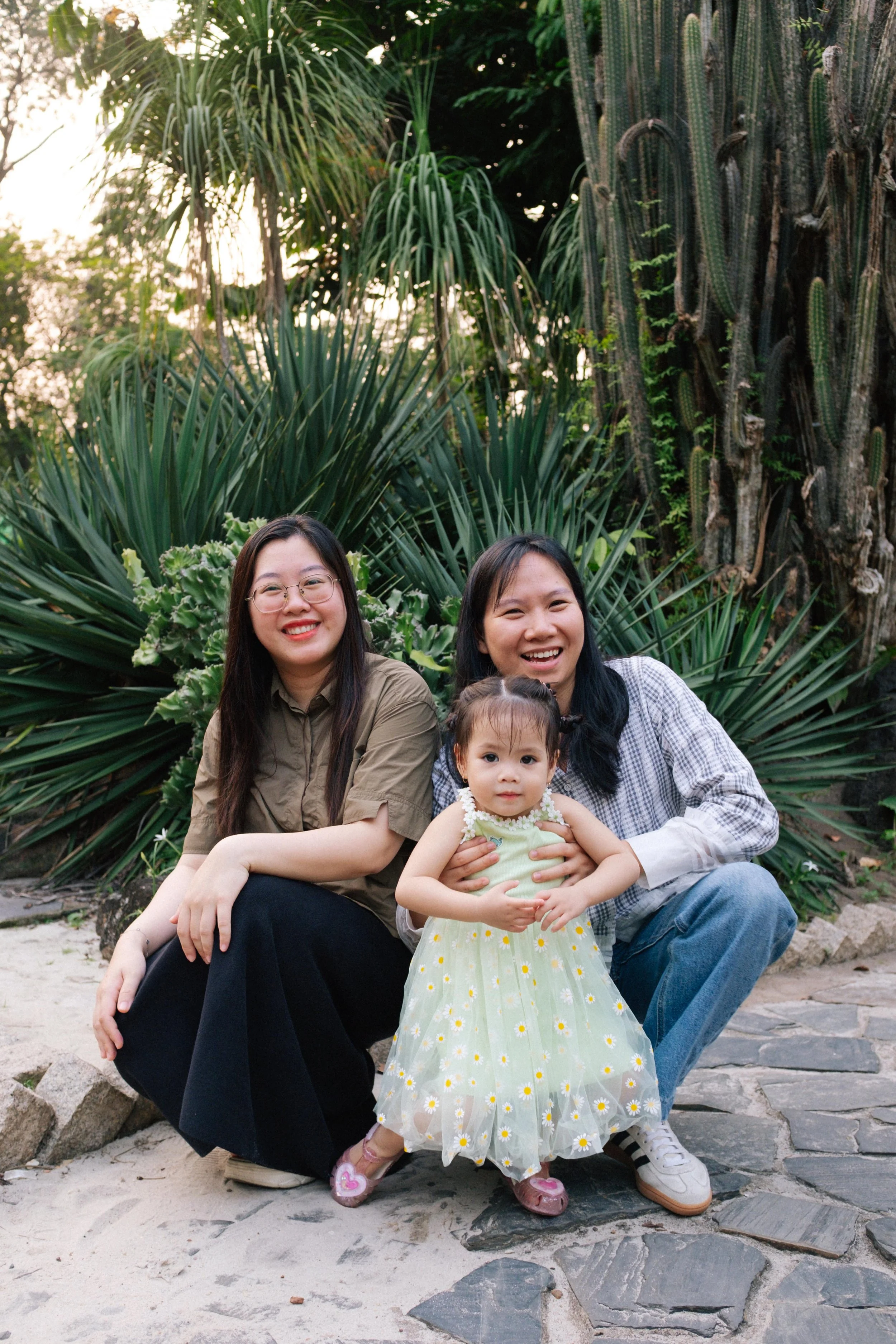 Two women and a young girl posing outdoors in front of lush green tropical plants, smiling at the camera.