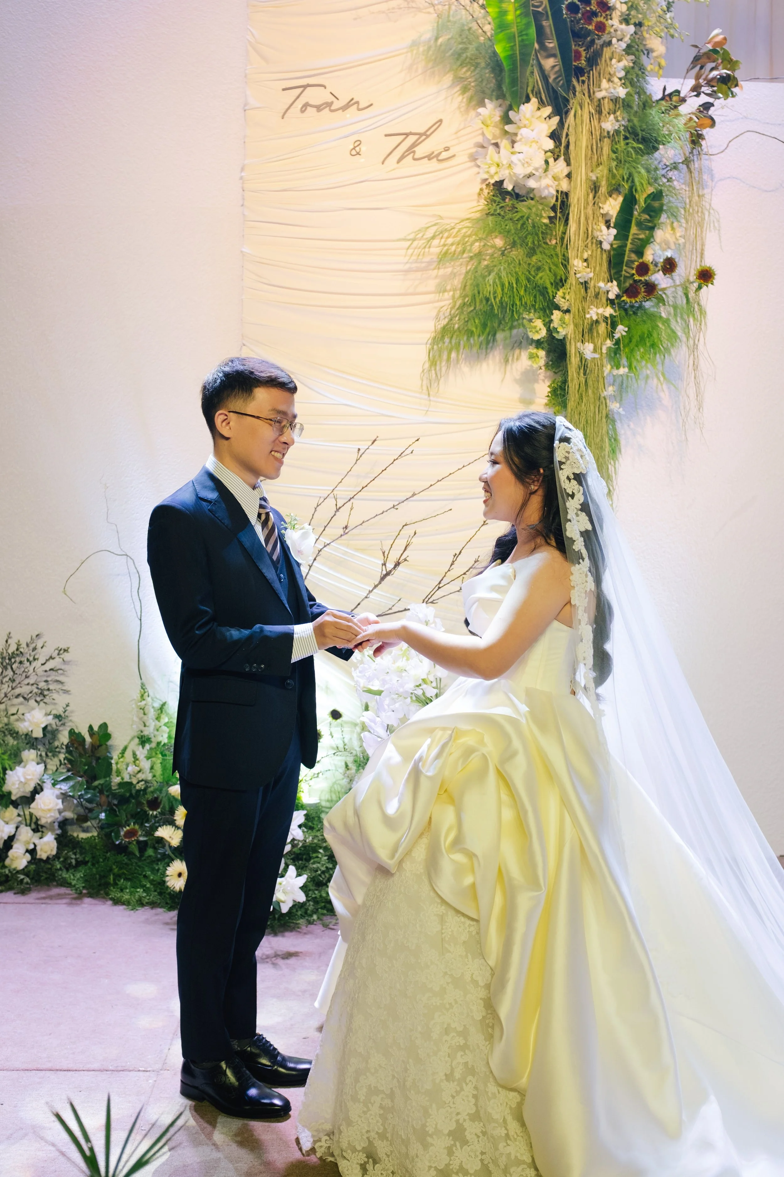 A bride and groom exchanging rings during a wedding ceremony, with a backdrop of flowers and greenery, and the words 'Toàn & Thư' on a draped fabric background.