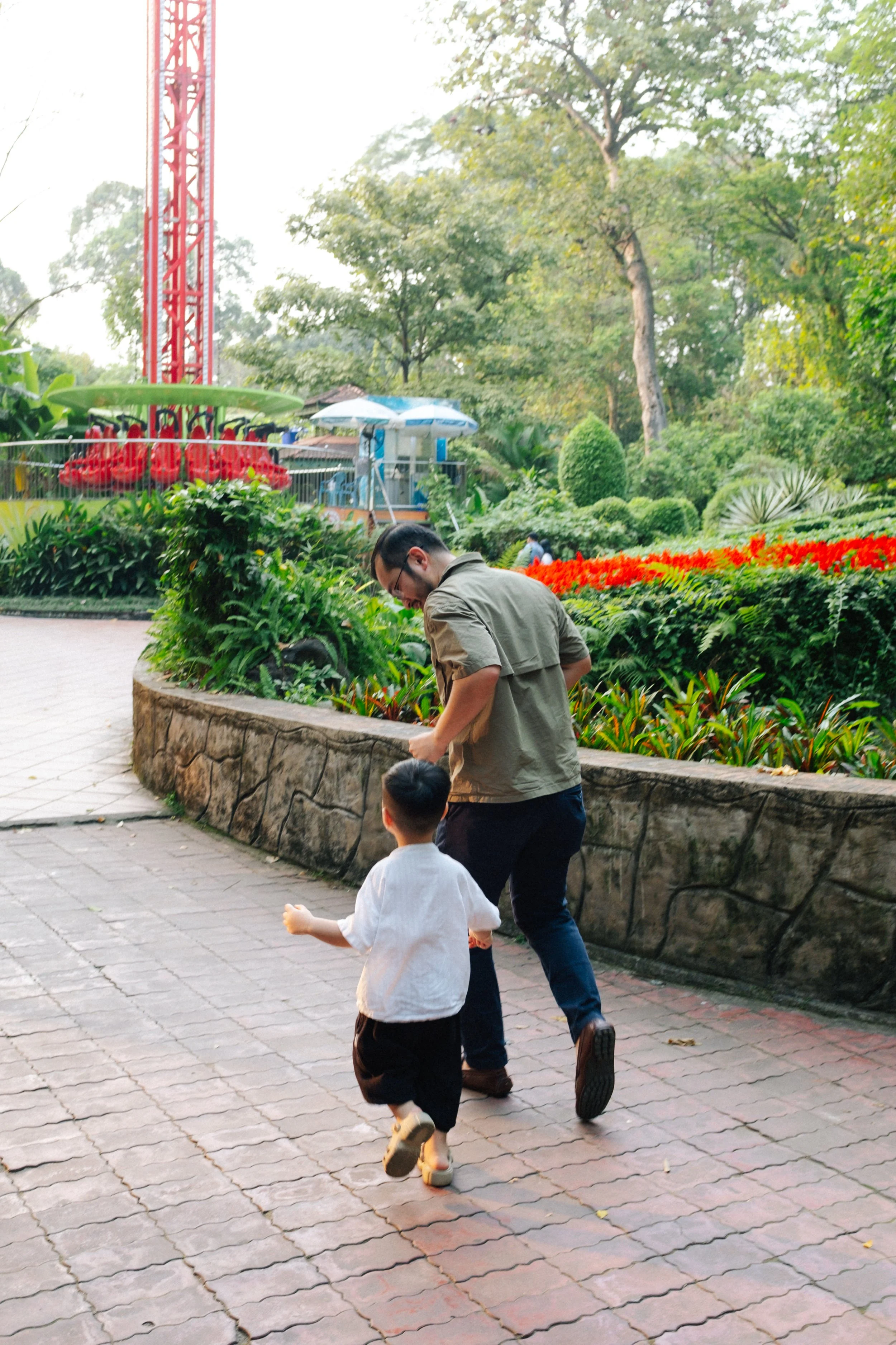 A man and a young boy walking along a paved pathway in a lush park, surrounded by green trees and colorful flowers.