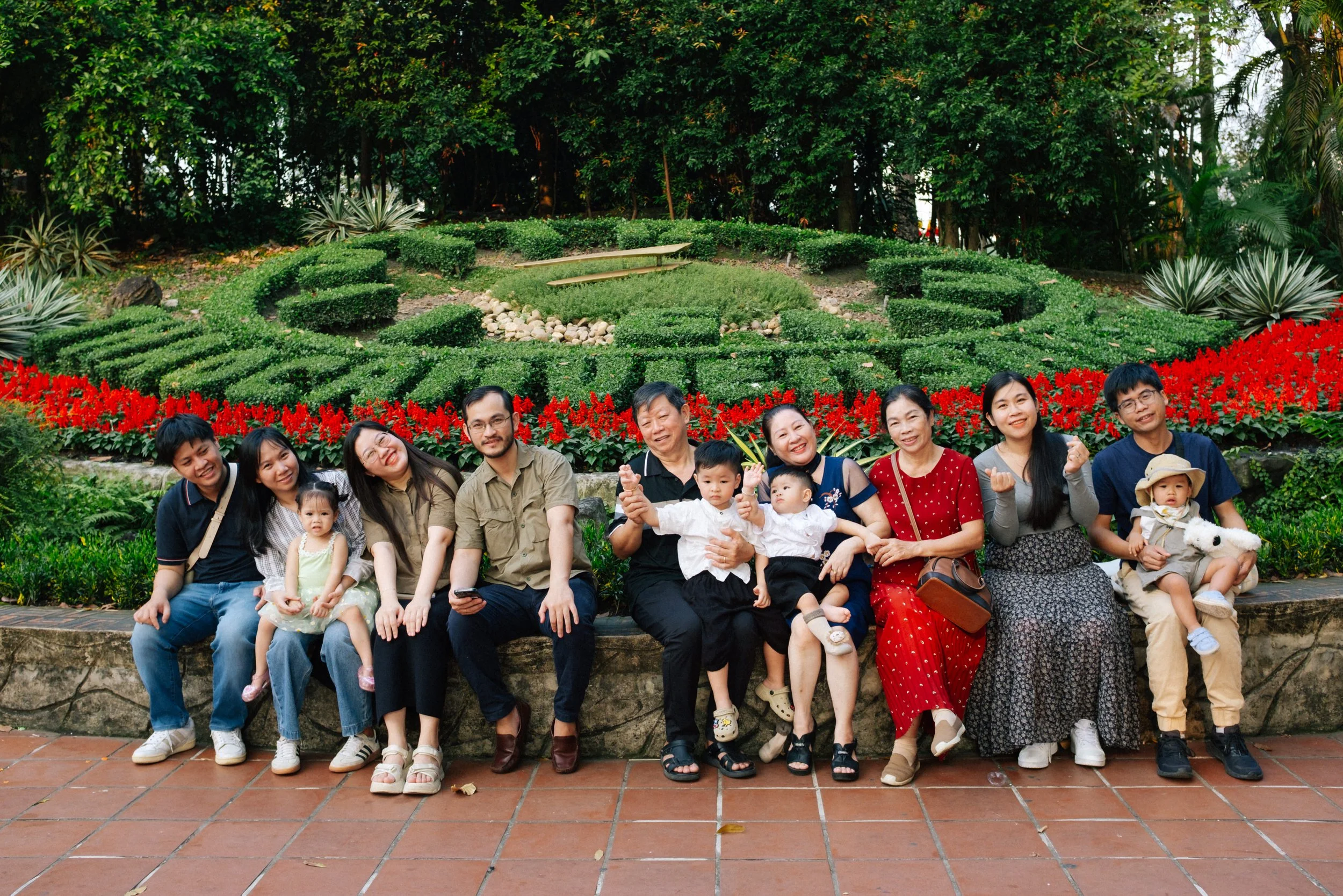 A group of 13 people, including children and adults, sitting on a stone ledge in front of a colorful flower garden with a clock shaped hedge in the background.