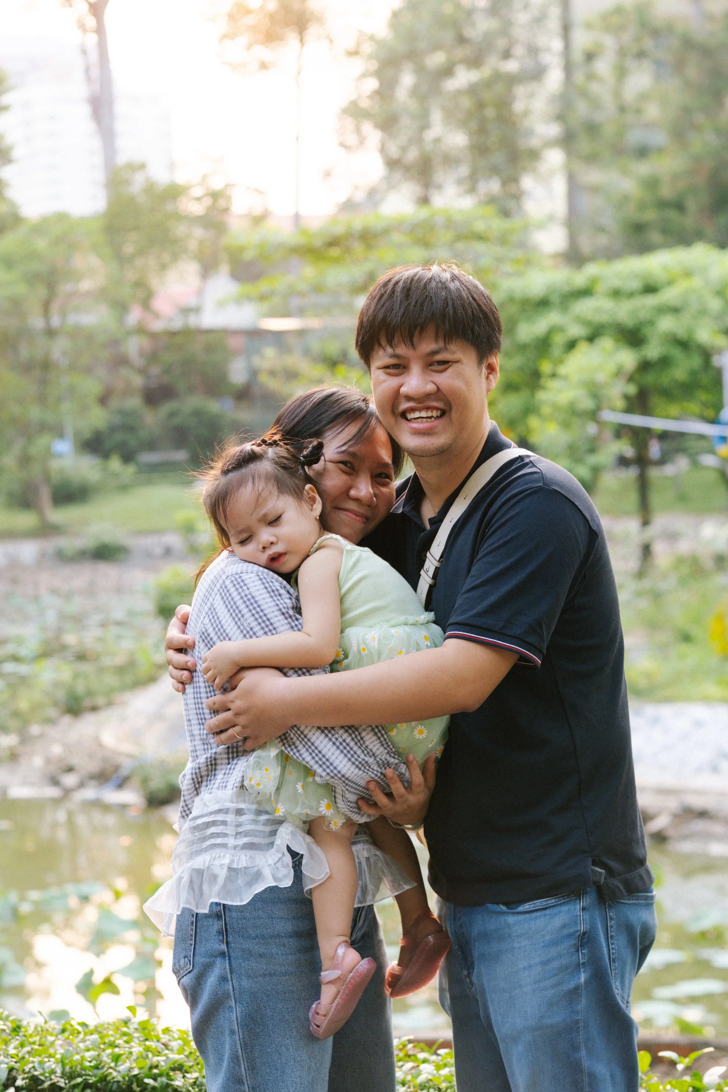 A happy family of three hugging outdoors near a pond with trees and buildings in the background, during daylight.