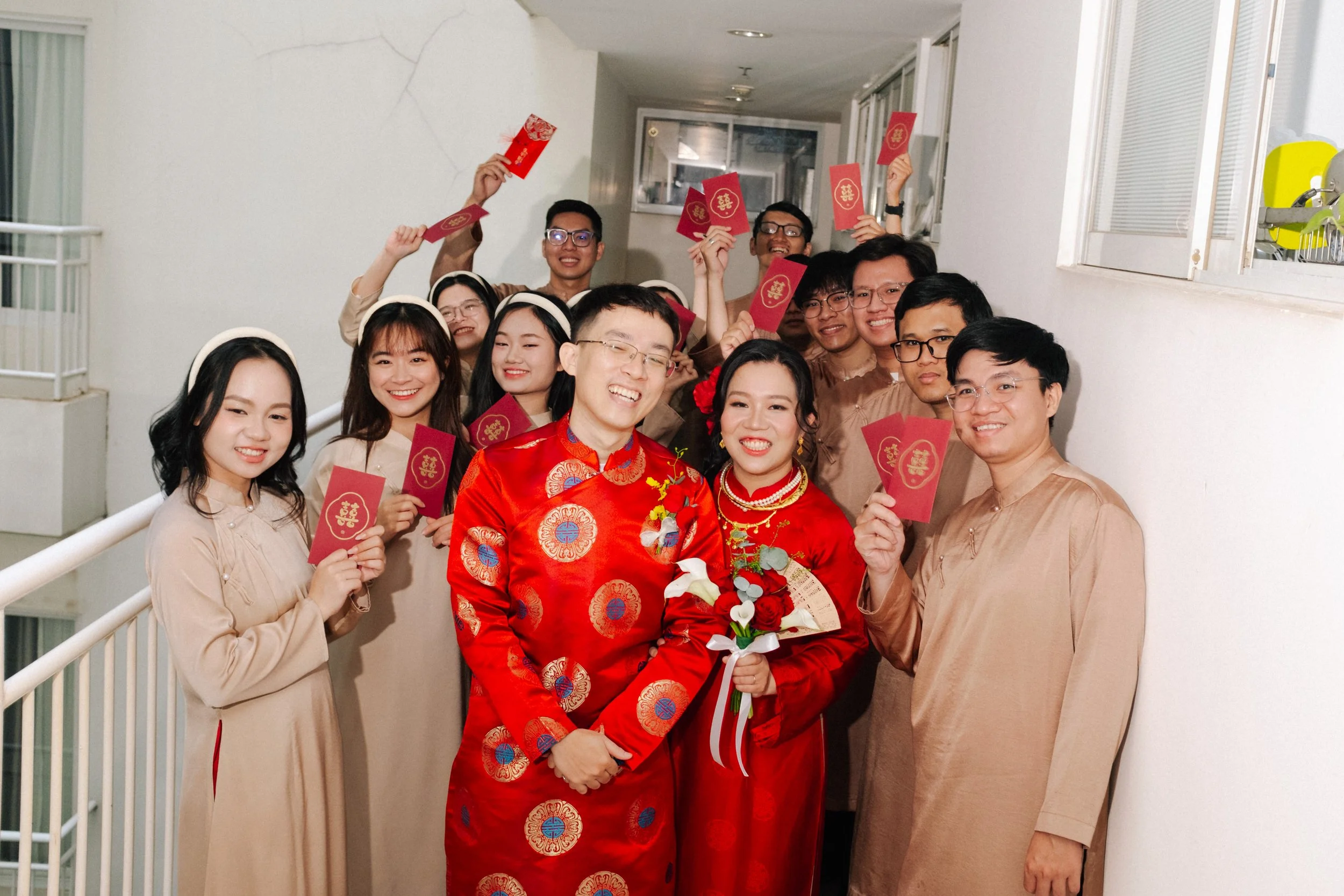 A group of people celebrating a wedding in traditional Asian attire, holding red envelopes with gold symbols, indoors near a white wall.