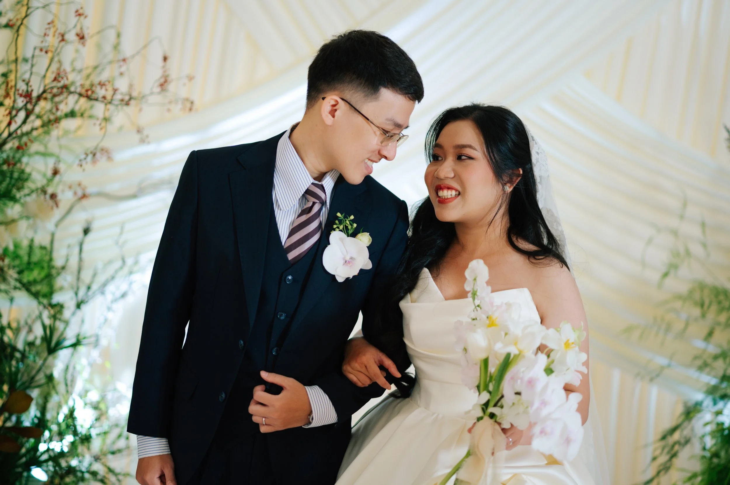 Bride and groom smiling closely, dressed in wedding attire, with flowers and floral decorations in the background.
