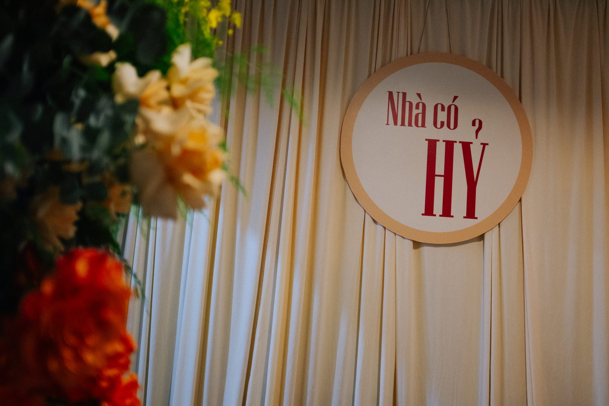 A round sign with Vietnamese text on a cream-colored curtain background, partially obscured by colorful flowers on the left.