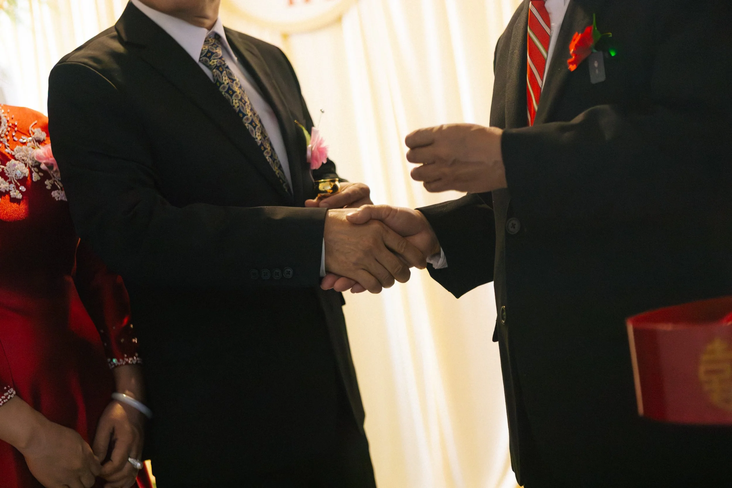Two men in formal attire shaking hands during a ceremony, with women in traditional clothing and floral decorations visible in the background.