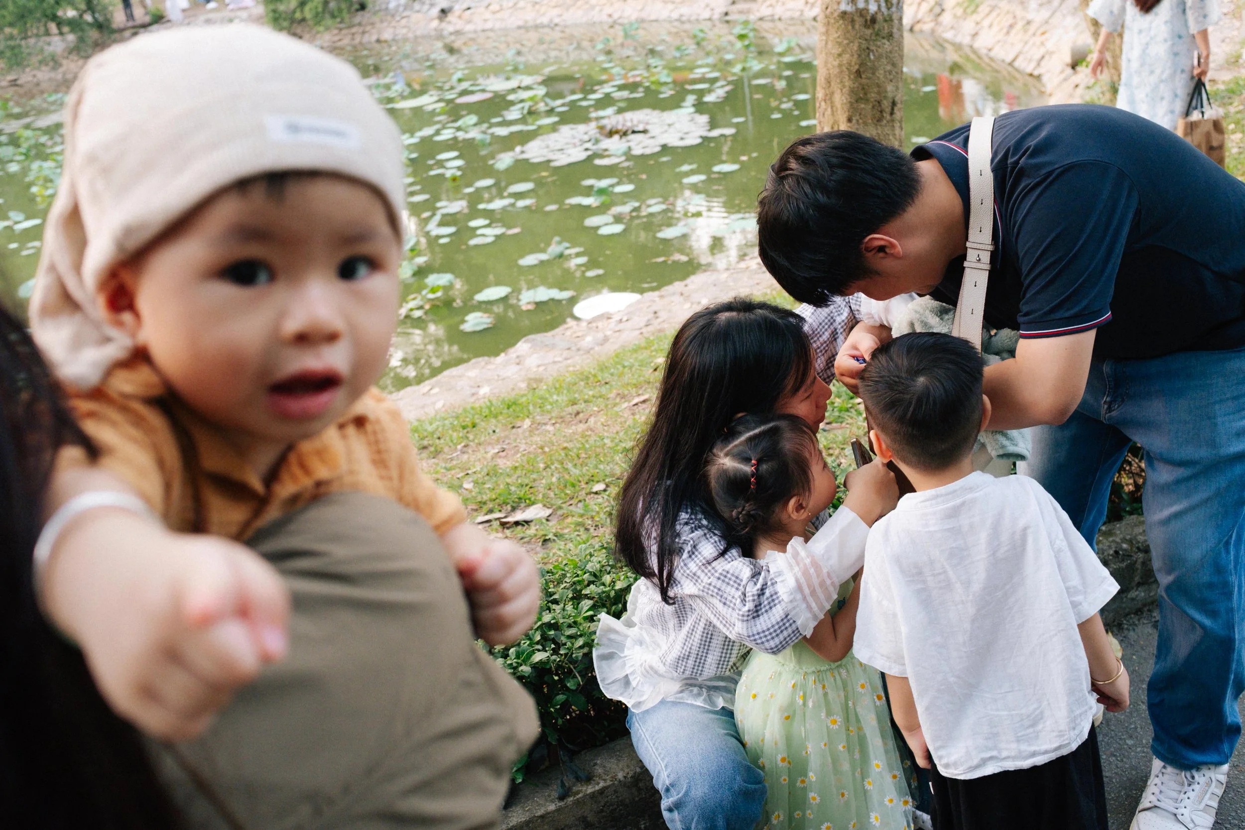 A young boy in a beige hat and brown shirt in the foreground, with a group of children and a woman near a pond in the background.