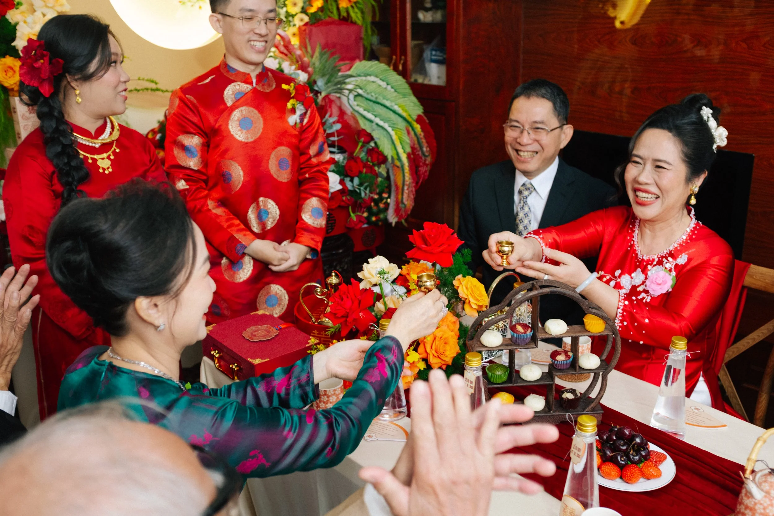 A joyful Chinese wedding banquet with women in traditional red dresses, a man in a red silk suit, and guests clinking teacups over a decorated table with flowers, cupcakes, and bottled drinks.