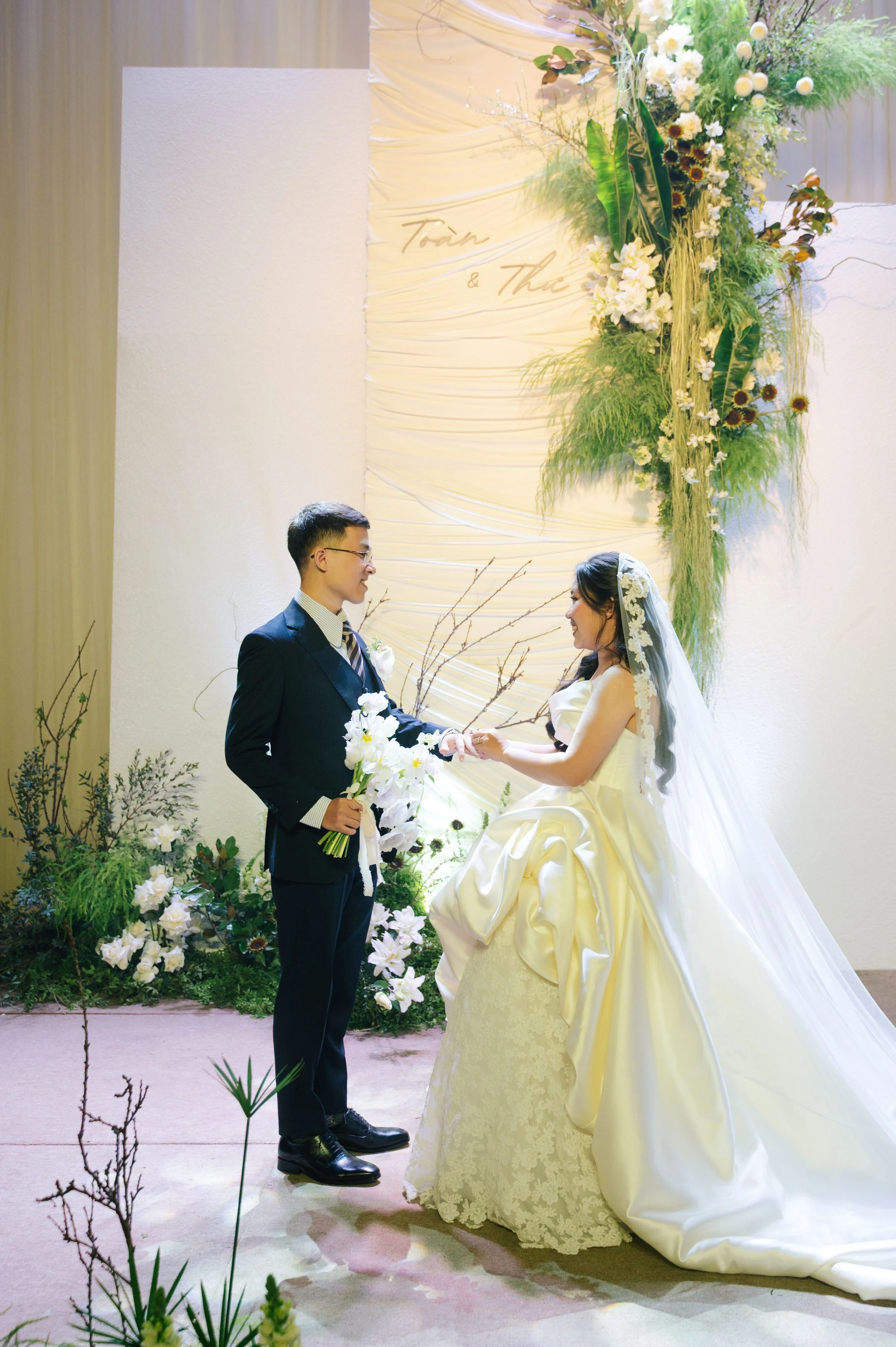 A bride and groom exchanging rings during their wedding ceremony, standing in front of a floral arrangement with white, green, and yellow flowers and a backdrop with gold lettering. The bride is wearing a white wedding gown with a long veil, and the 
