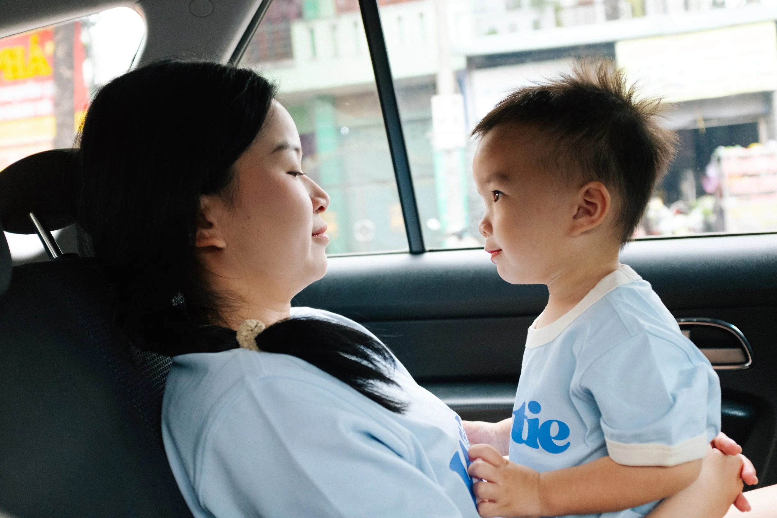 A woman sitting in the backseat of a car with a young boy standing in front of her, holding her hands and looking into her eyes.