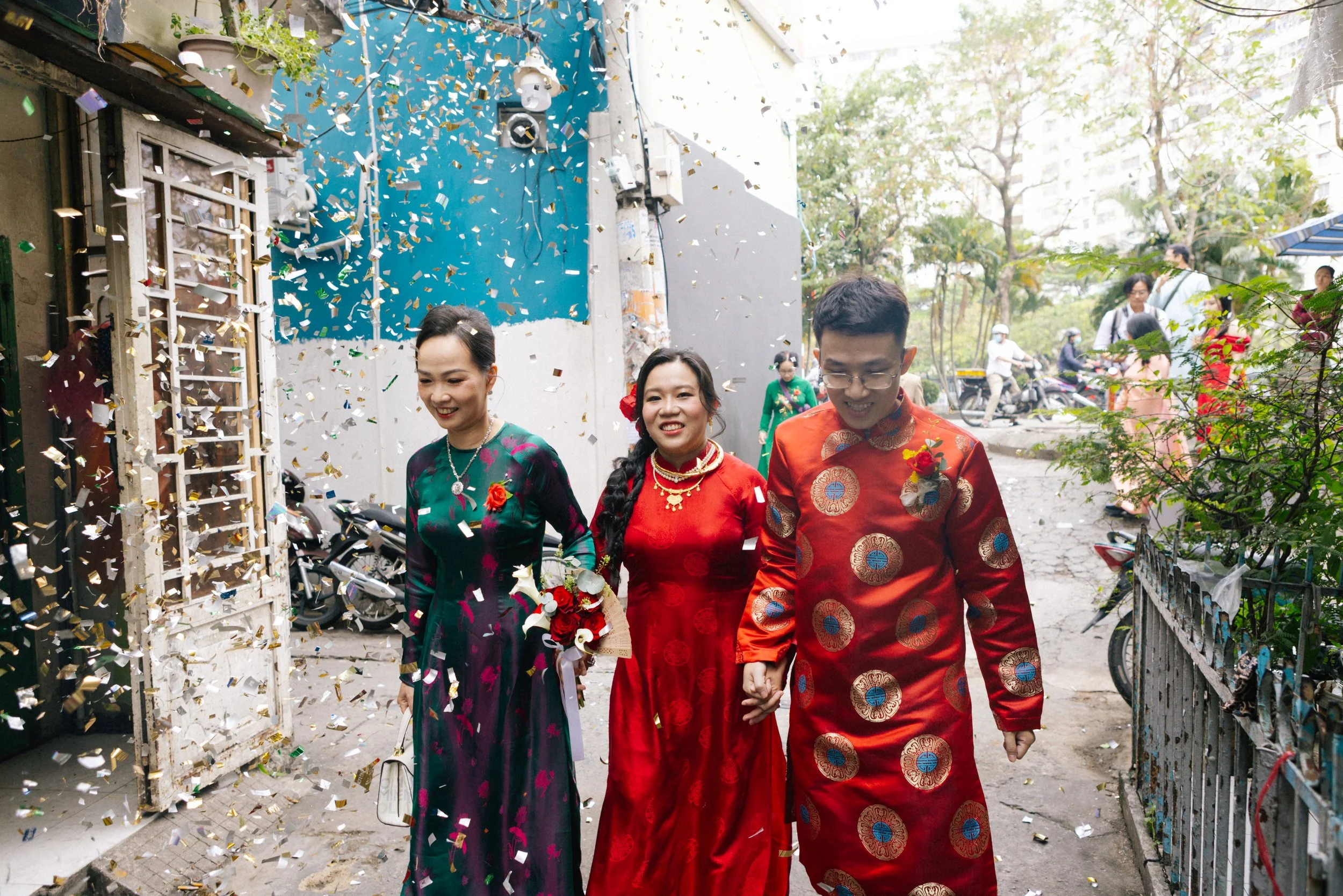 Three people in traditional Vietnamese clothing walking through a confetti celebration outside with trees and motorcycles in the background.