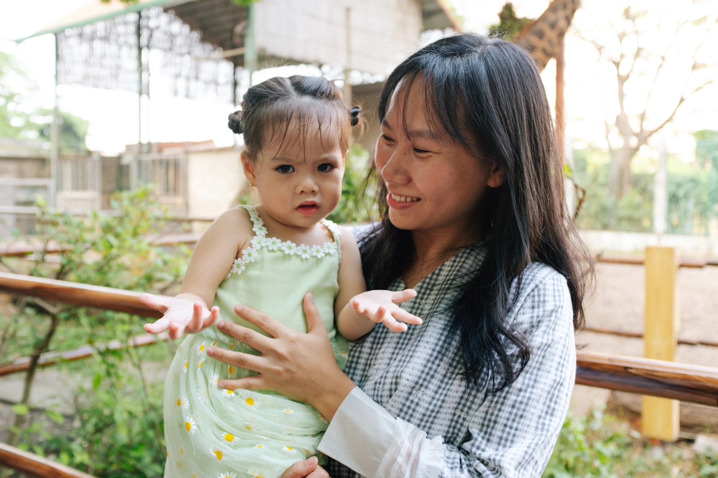 A woman holding a young girl outdoors, with trees and a wooden fence in the background.