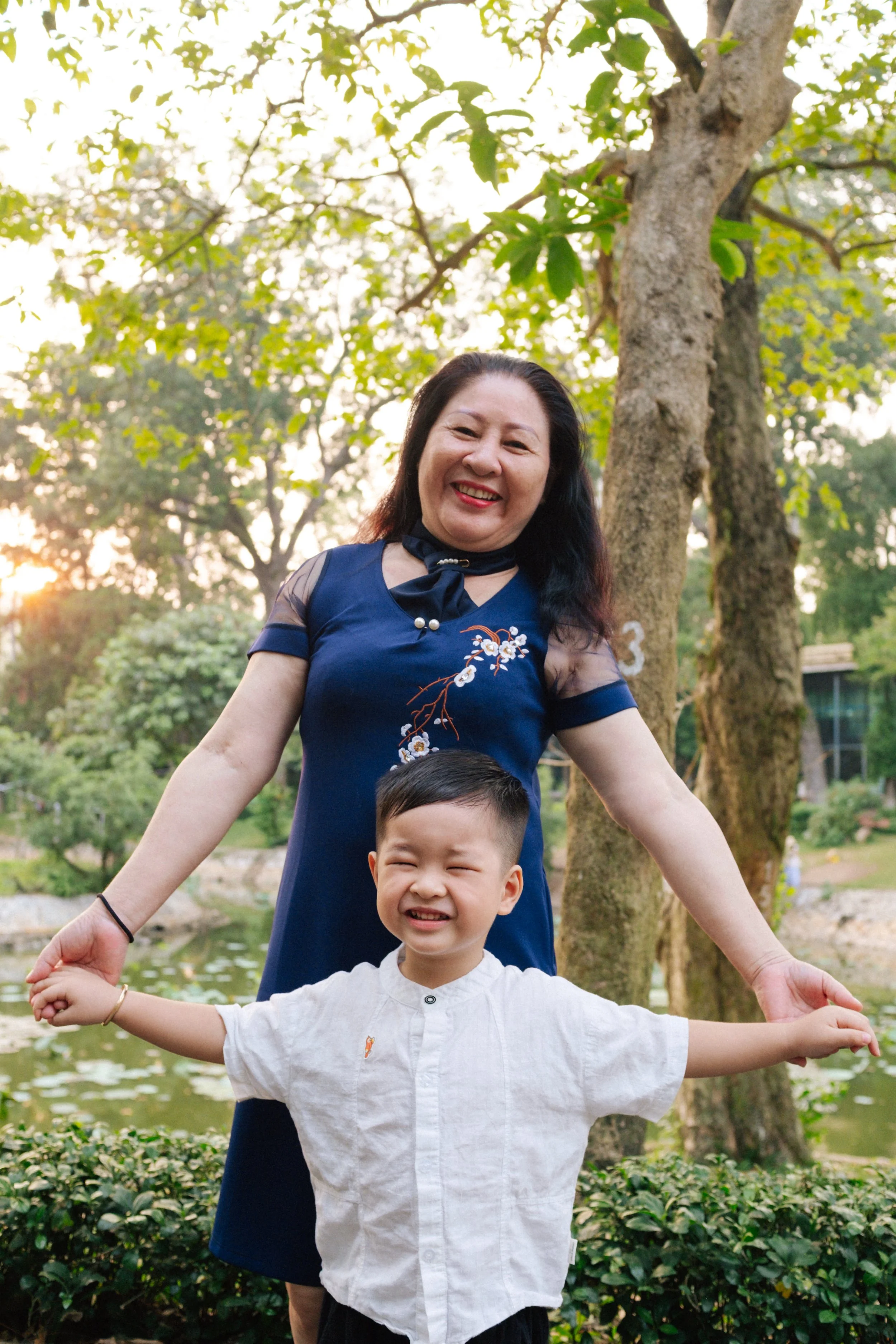 A smiling woman and young boy holding hands outdoors near a pond with trees and greenery in the background, during sunset.
