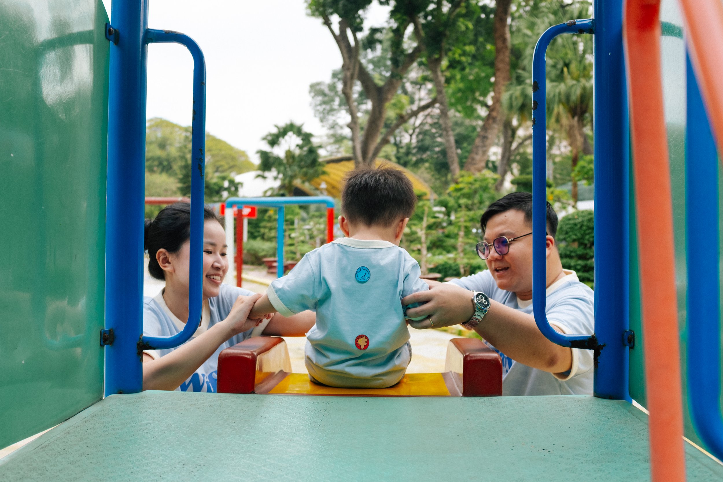 Two children and two adults playing on a playground slide outdoors, surrounded by trees.