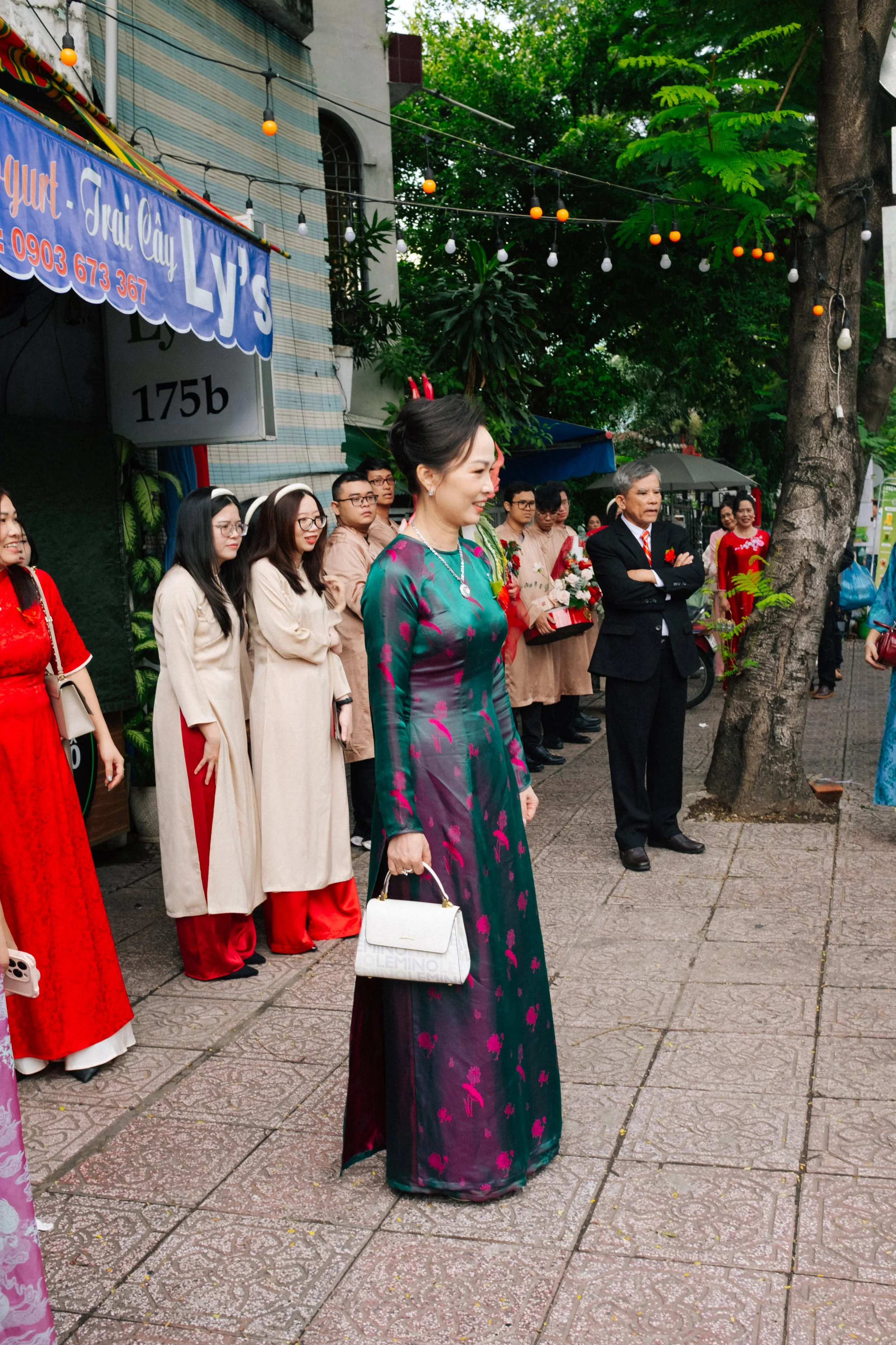 A group of people standing outdoors on a sidewalk during a celebration or event, some holding flowers, with colorful traditional clothing and a woman in a floral dress in the foreground. There are trees, string lights, and a sign for LV's Yogurt Tray