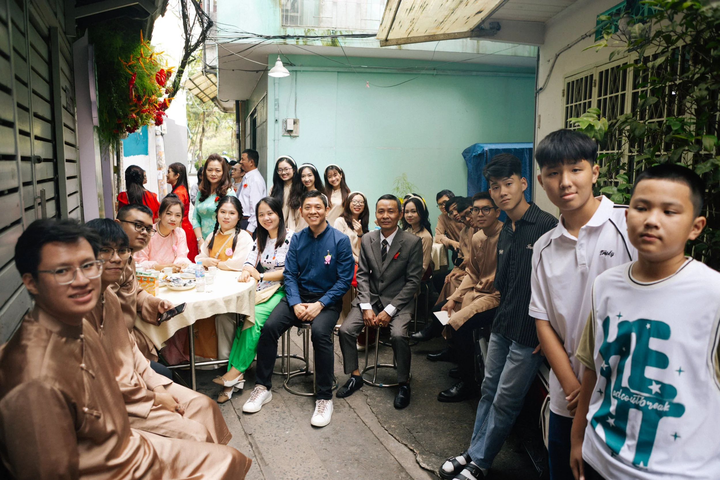A large group of people gathered in an alley for a celebration, sitting and standing around a table with drinks and snacks, smiling at the camera.