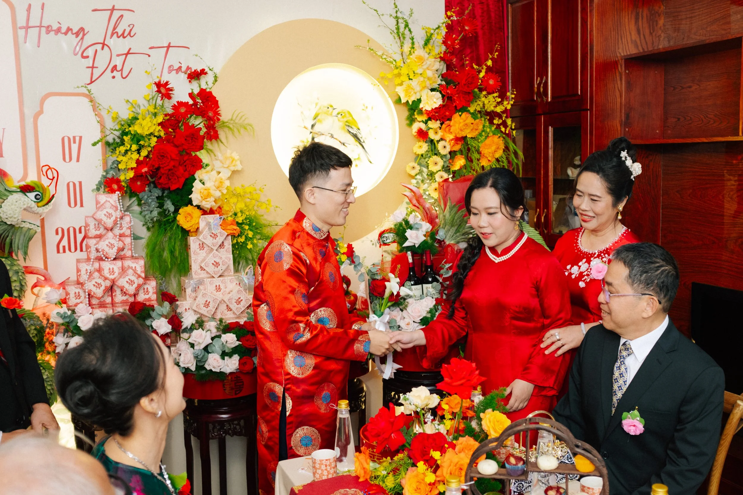 A traditional Vietnamese wedding celebration with a couple exchanging rings at the altar, surrounded by family members, vibrant red and yellow floral decorations, and festive symbols.