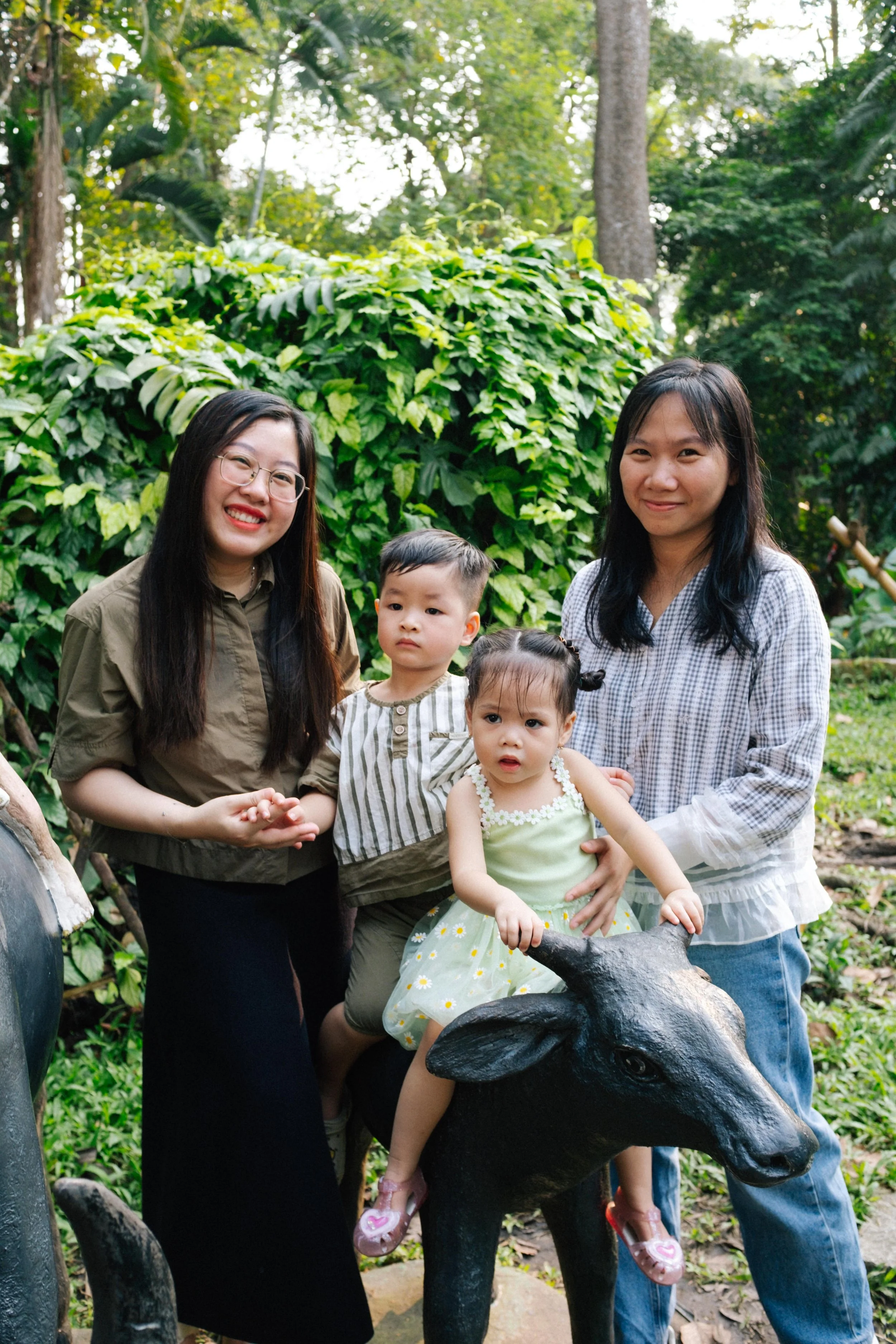 Two women and two children standing outdoors with greenery in the background. One child is sitting on a black animal sculpture, with a girl in a light green dress holding the sculpture. The girls and boys are dressed casually and smiling at the camer