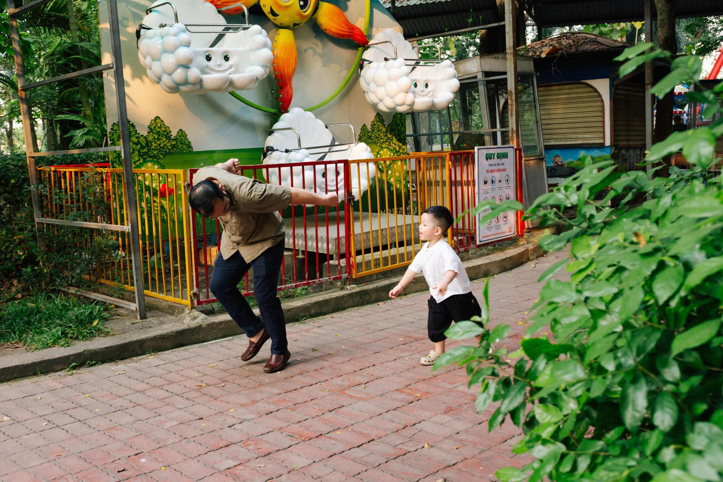 A woman and a young boy playing outside near a colorful amusement ride with cloud and sun decorations, surrounded by green foliage and trees.