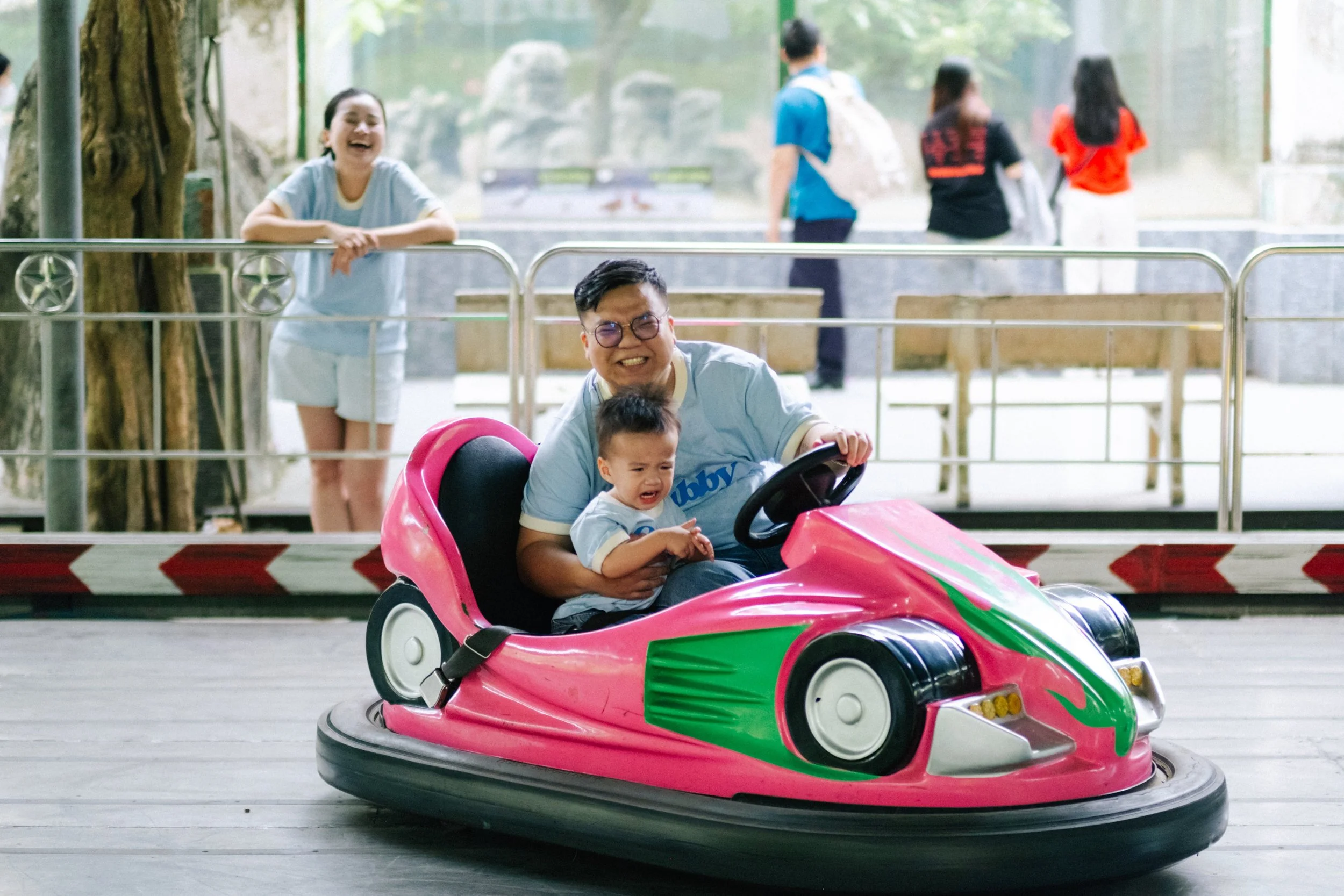 A man and a young boy riding a pink bumper car at an amusement park. The boy appears to be crying or upset, while the man is smiling. There are two women in the background near a railing, smiling and watching the ride, with other park visitors visibl