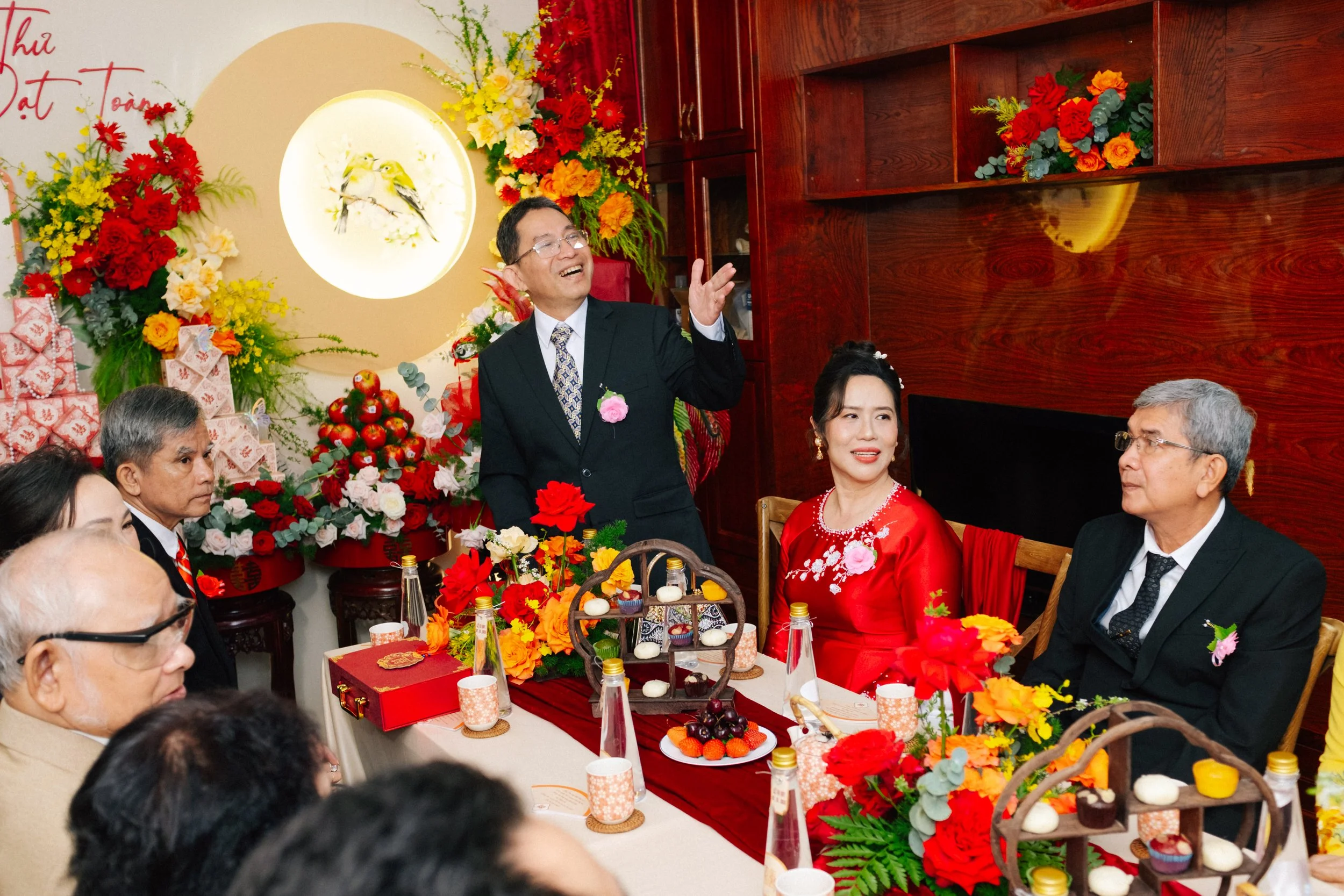 A group of people at a wedding celebration, with a man standing and speaking, a woman in a red dress sitting at the table, and several other guests seated. The table is decorated with colorful flowers, food, and drinks. The background features a wall