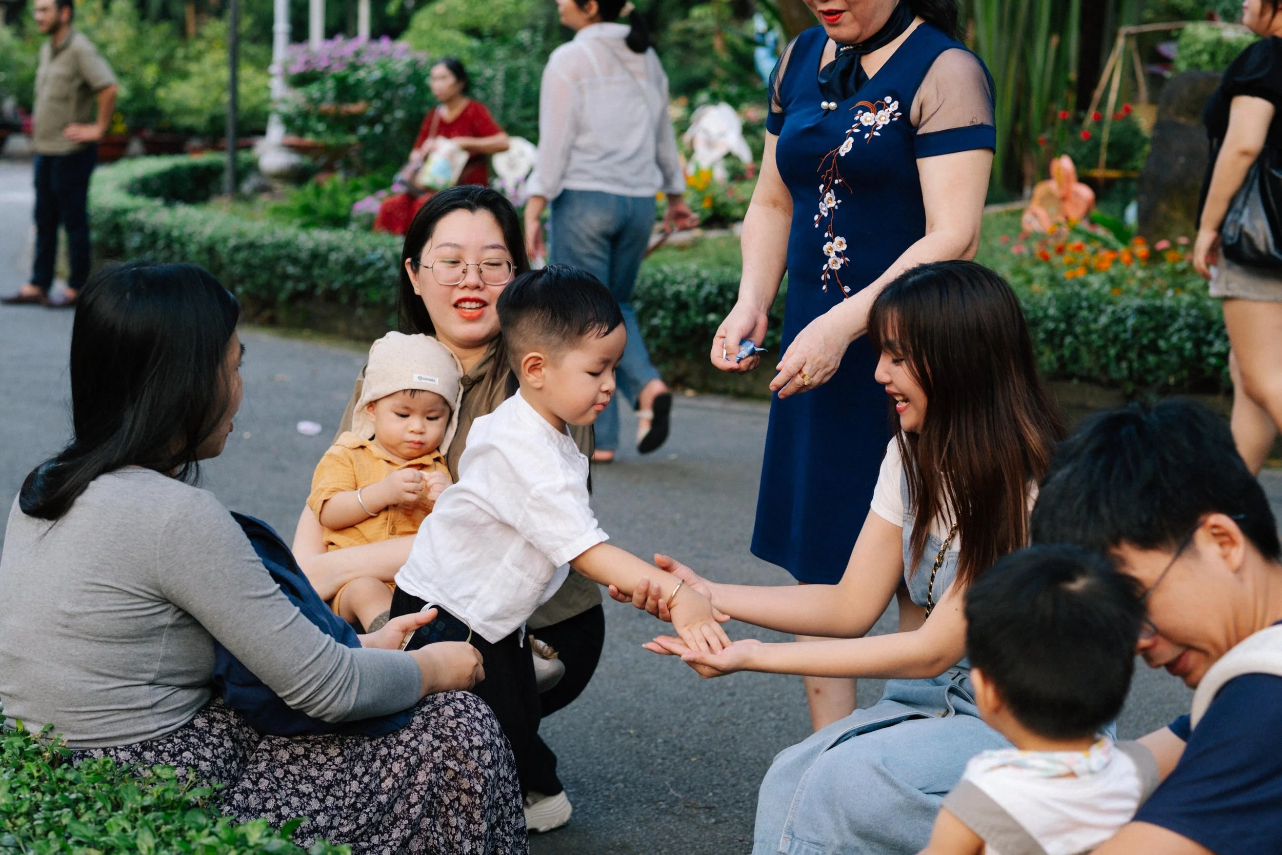 People gathered in a park, with a woman in a blue dress handing a young child a small object. The child reaches towards a woman sitting on the ground, who is smiling. Other children and adults are nearby, with greenery and flowers in the background.