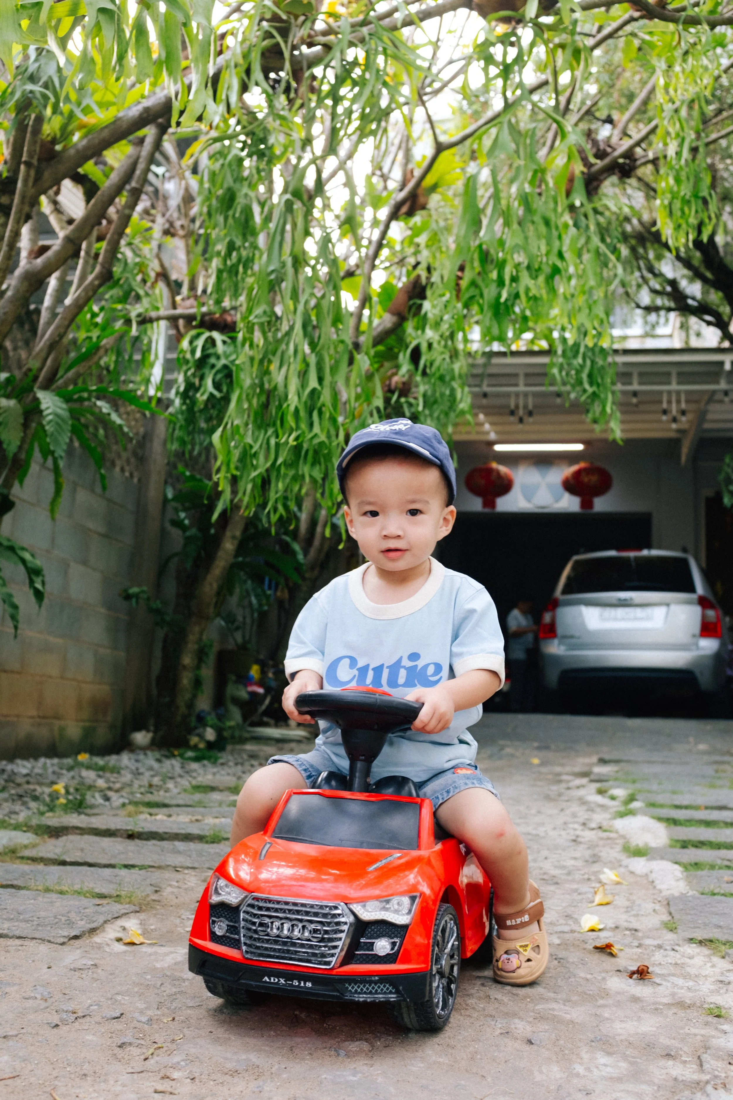 Young boy sitting on a red toy car outdoors with trees and a parked silver SUV in the background.