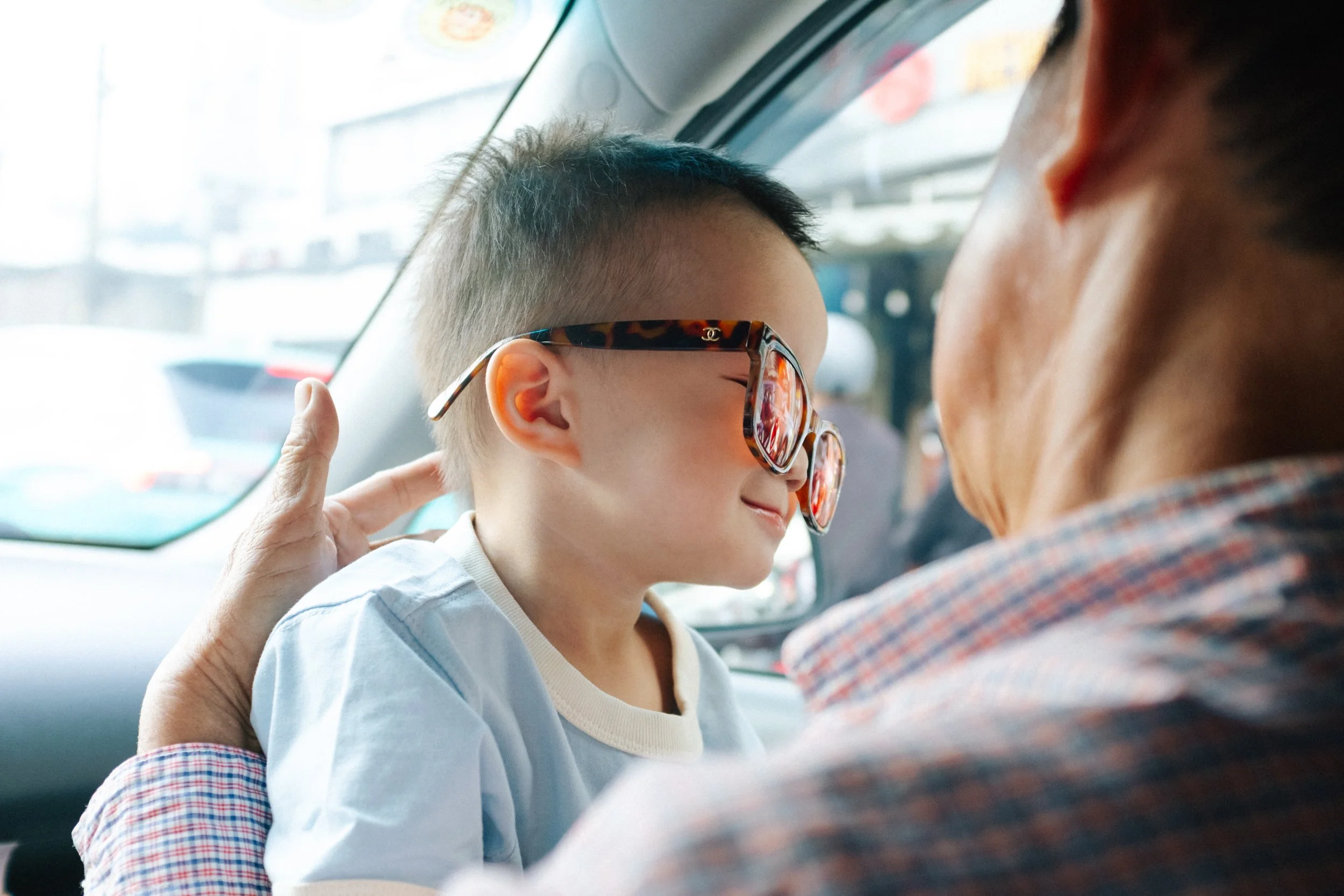 A young boy wearing large, branded sunglasses is leaning into an older person, possibly a grandmother, in the backseat of a car. The older person is gently touching the boy's head, and they appear to be sharing a tender moment.