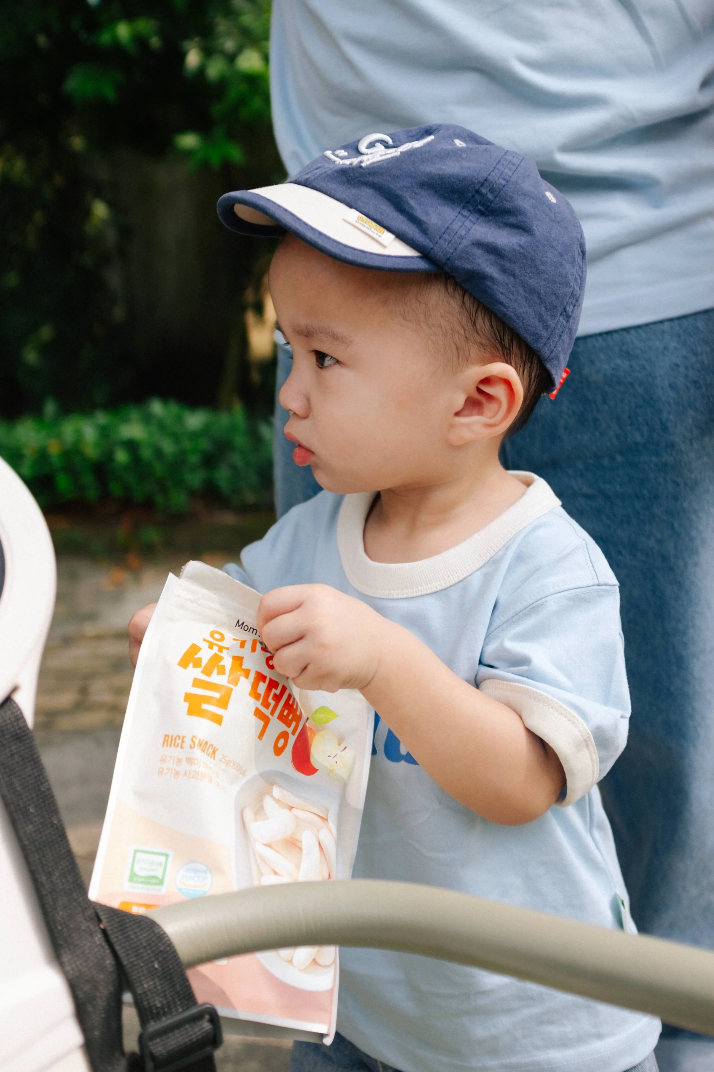 Young boy wearing a blue cap and a light blue shirt holding a rice snack packet outdoors in a park.