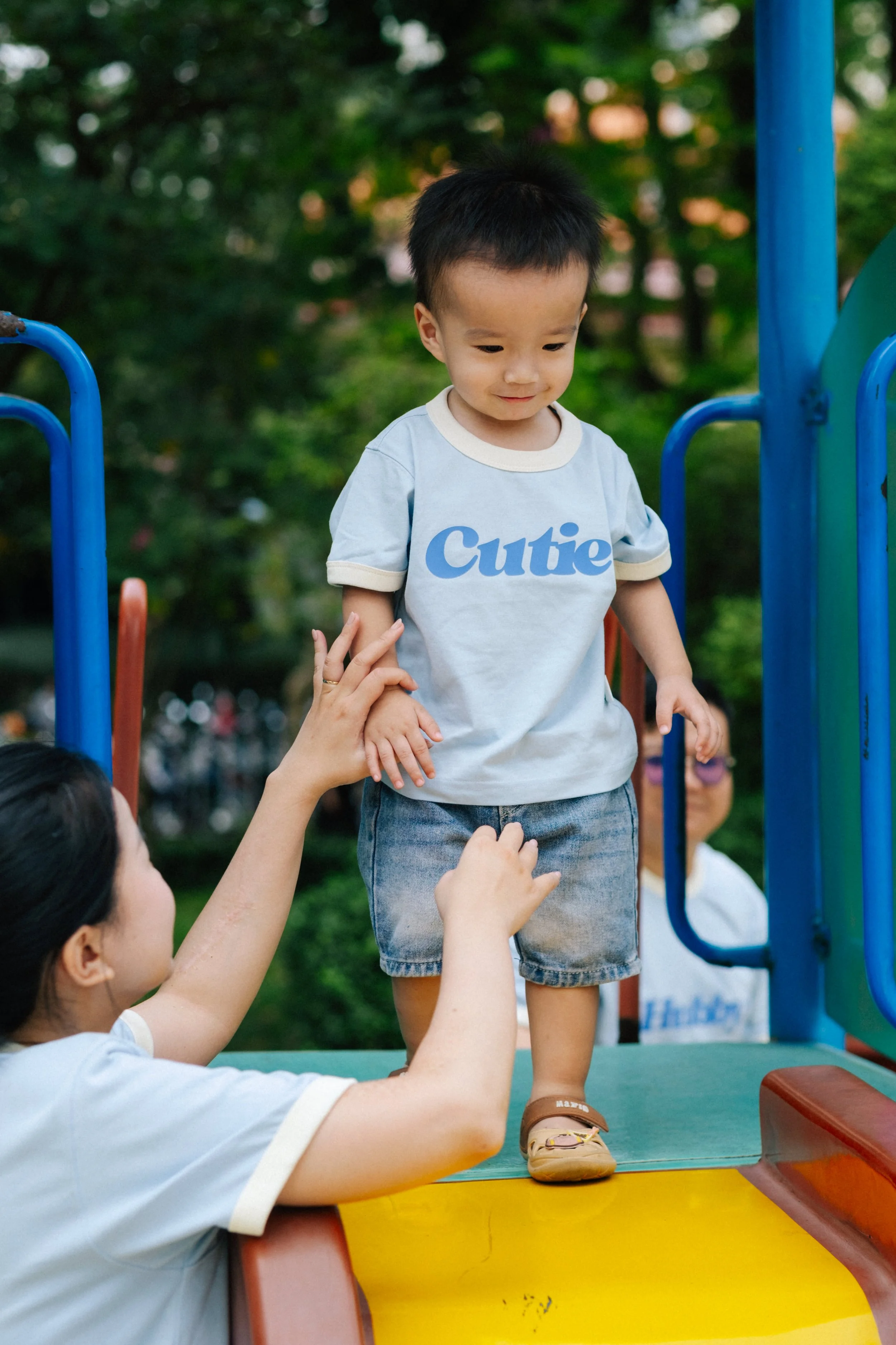 A young child standing on a playground structure, being supported by a woman, with trees and other people in the background.
