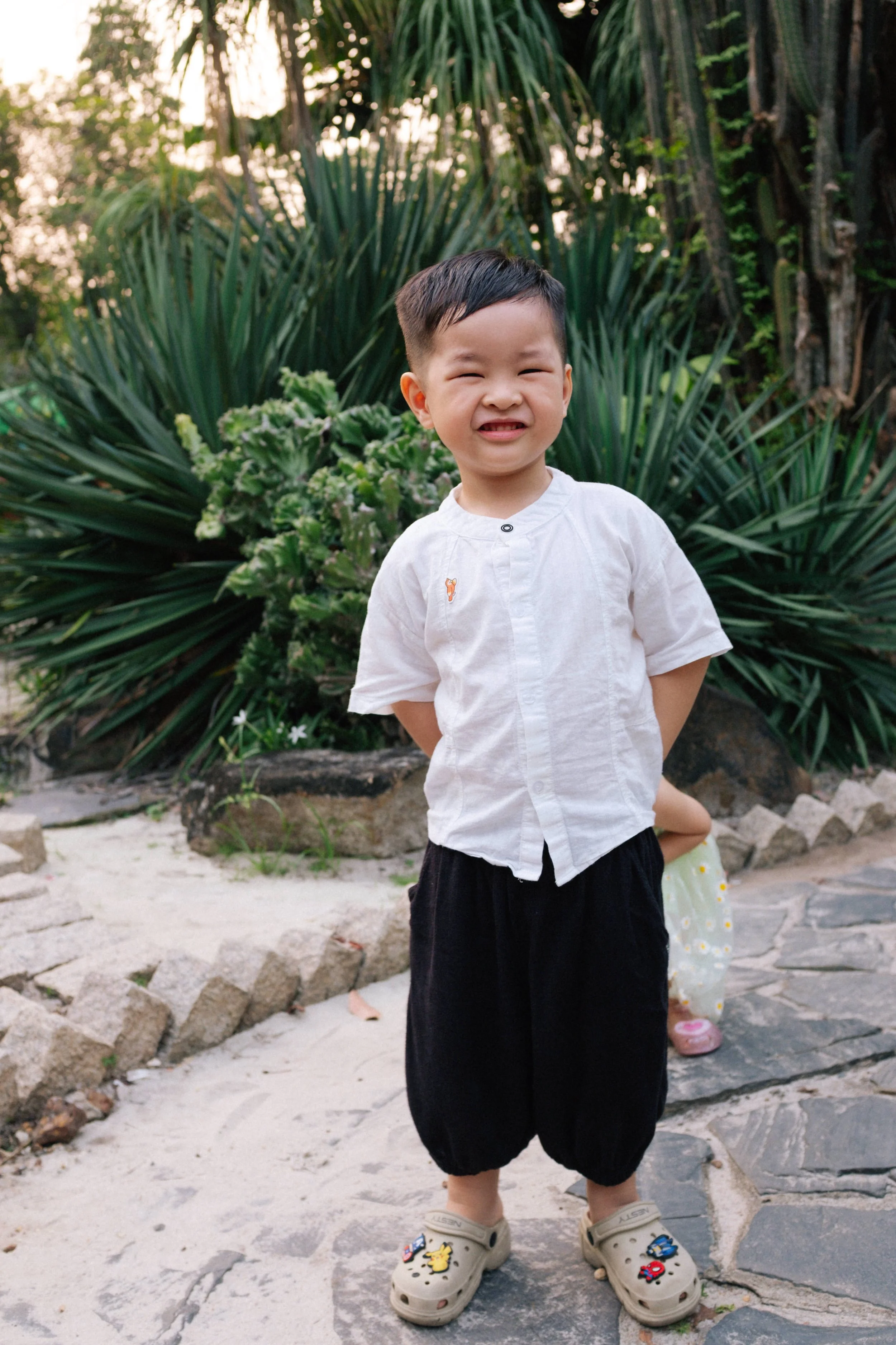 A young boy smiling outdoors, standing on a stone pathway with lush greenery behind him.