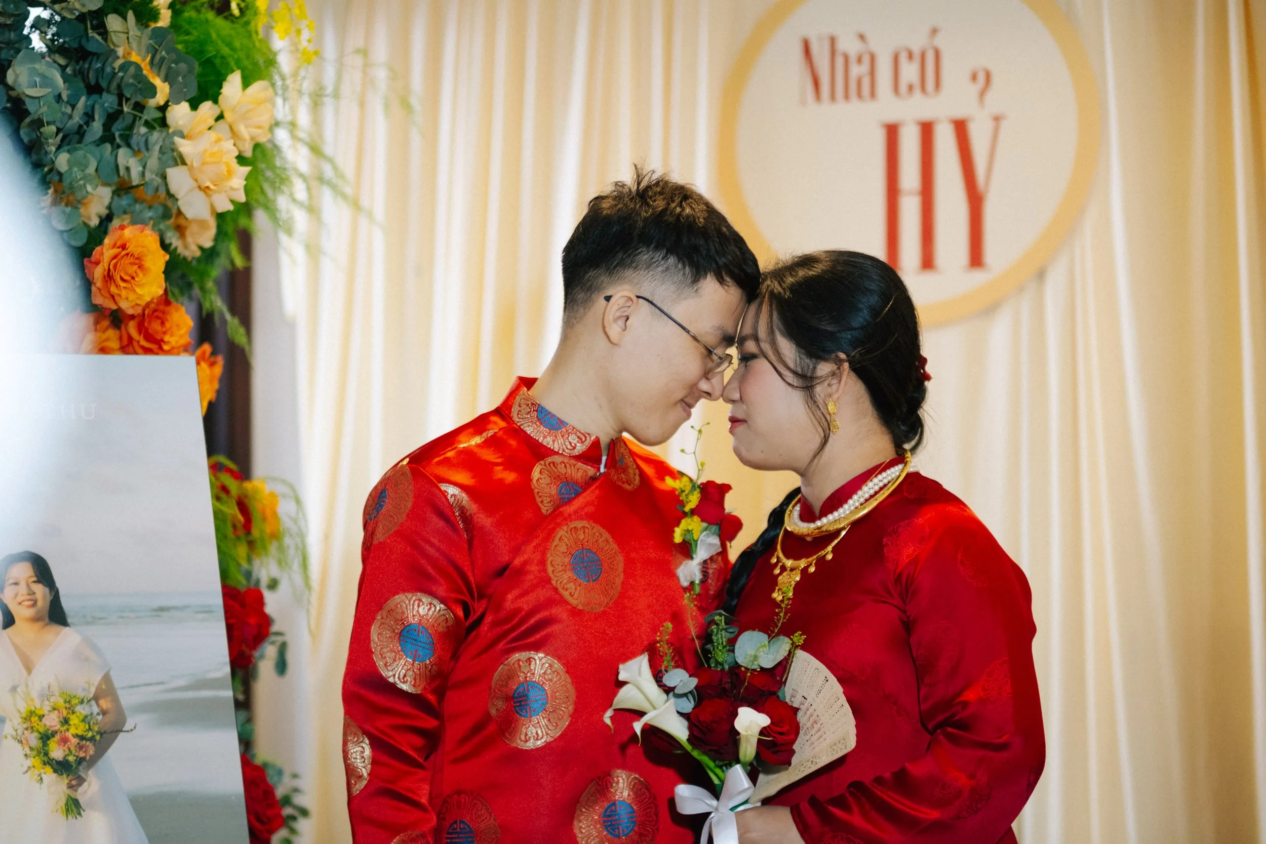 A couple dressed in traditional red Vietnamese wedding attire, standing close with foreheads touching, holding a bouquet of flowers, during their wedding ceremony indoors with floral decorations and a sign in Vietnamese in the background.