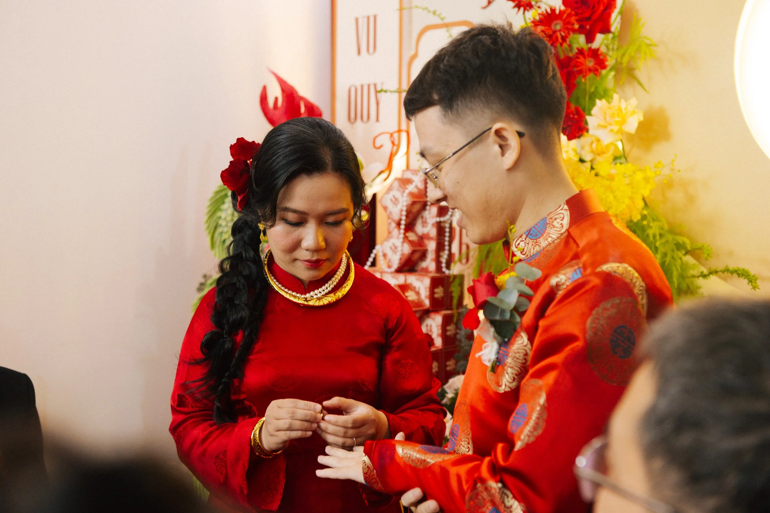 A couple dressed in traditional Chinese attire exchanging rings during a wedding ceremony, with festive red and gold decorations and flowers in the background.