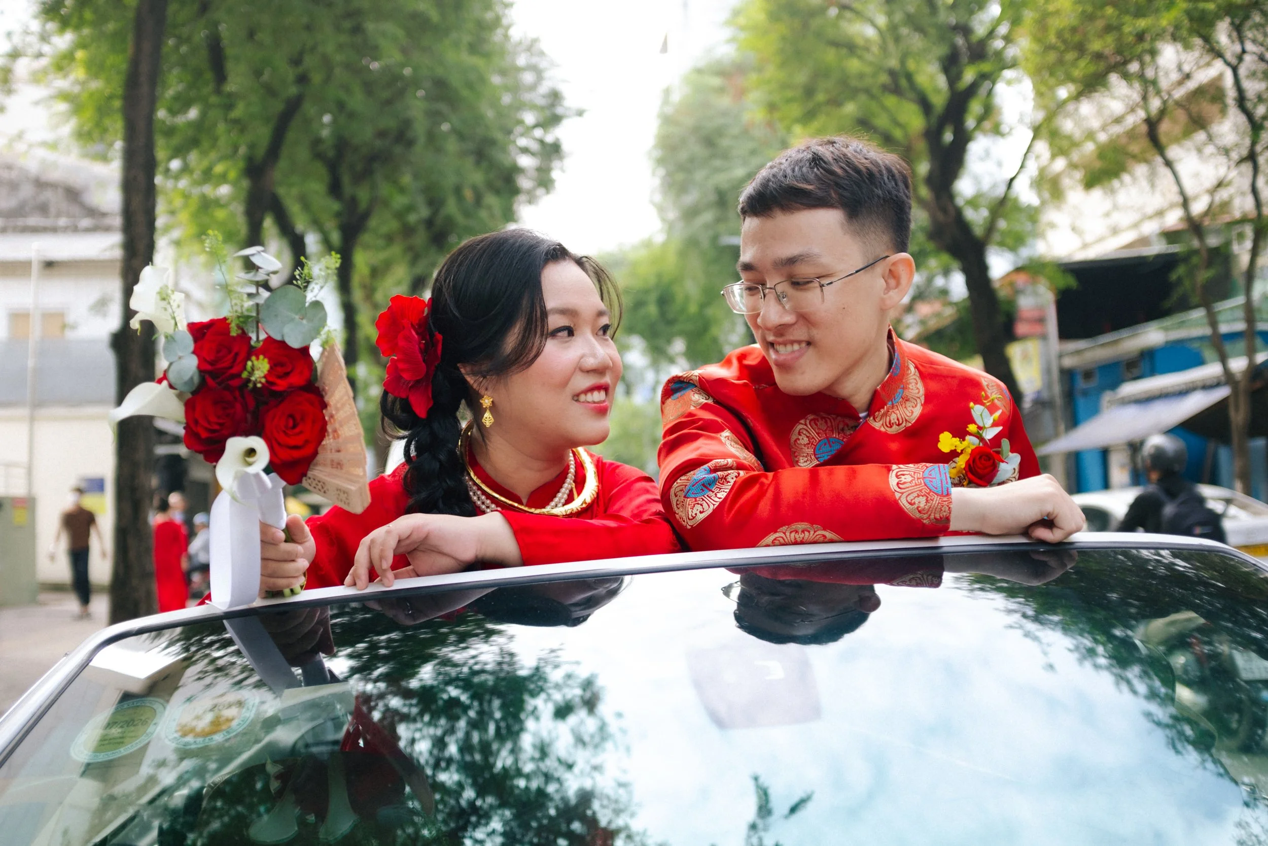 A newly married couple in traditional red Chinese wedding attire smiling at each other, sitting in a car with a bouquet of red roses and white flowers, on a tree-lined street.