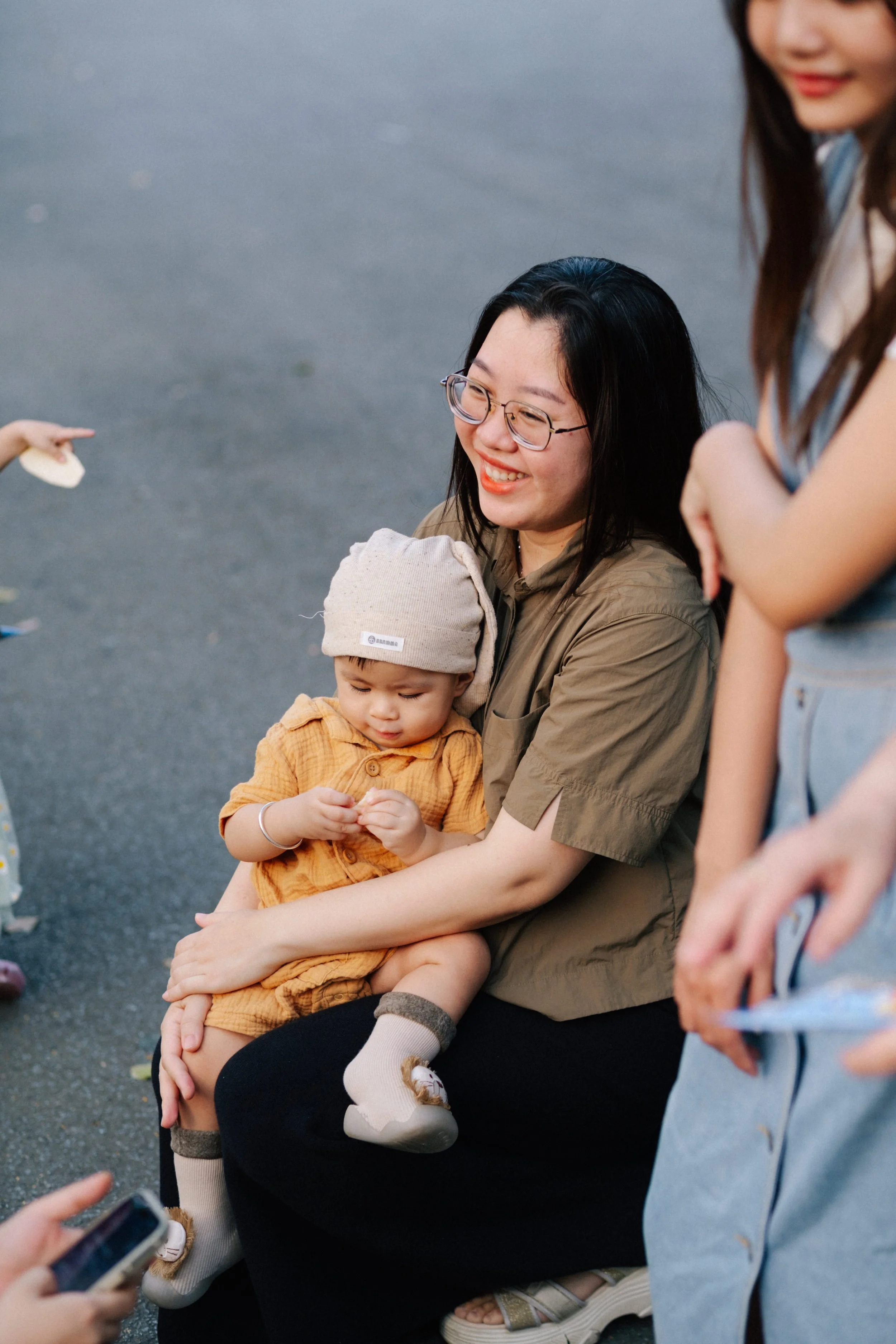 A woman with glasses holding a young child in a yellow outfit and beige hat, sitting on her lap on a gray pavement, surrounded by other people, smiling and engaging with each other.