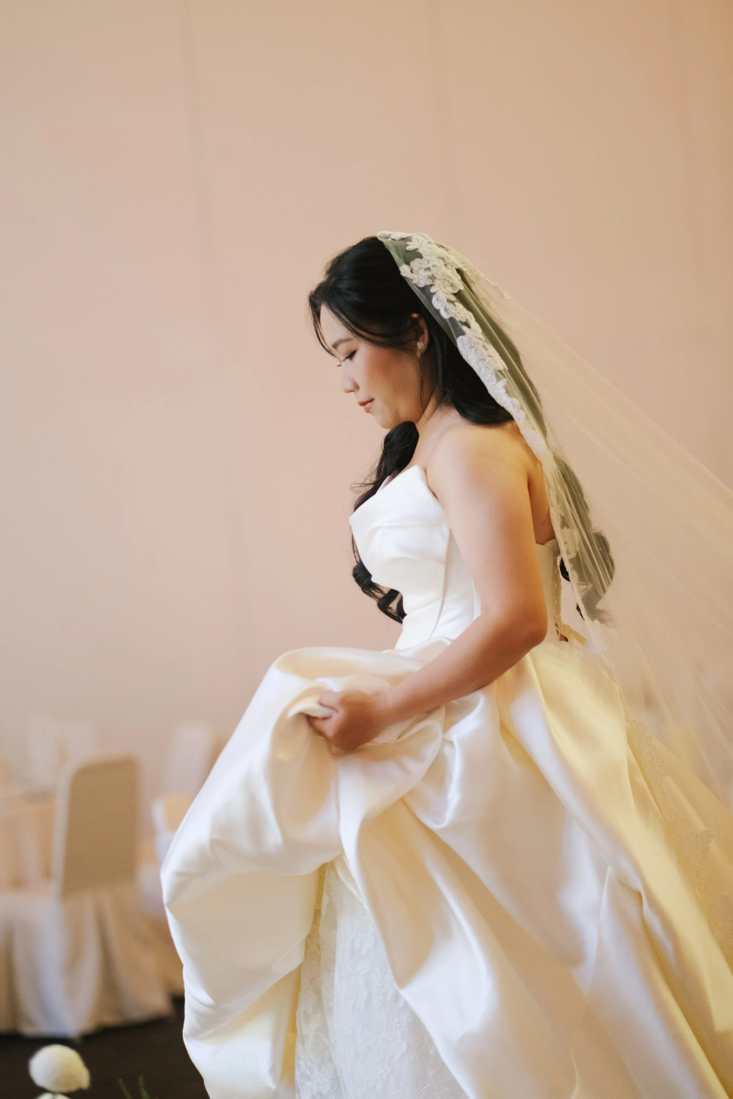 A bride in a white wedding dress, holding up her dress with one hand and wearing a wedding veil, standing in a softly lit room.