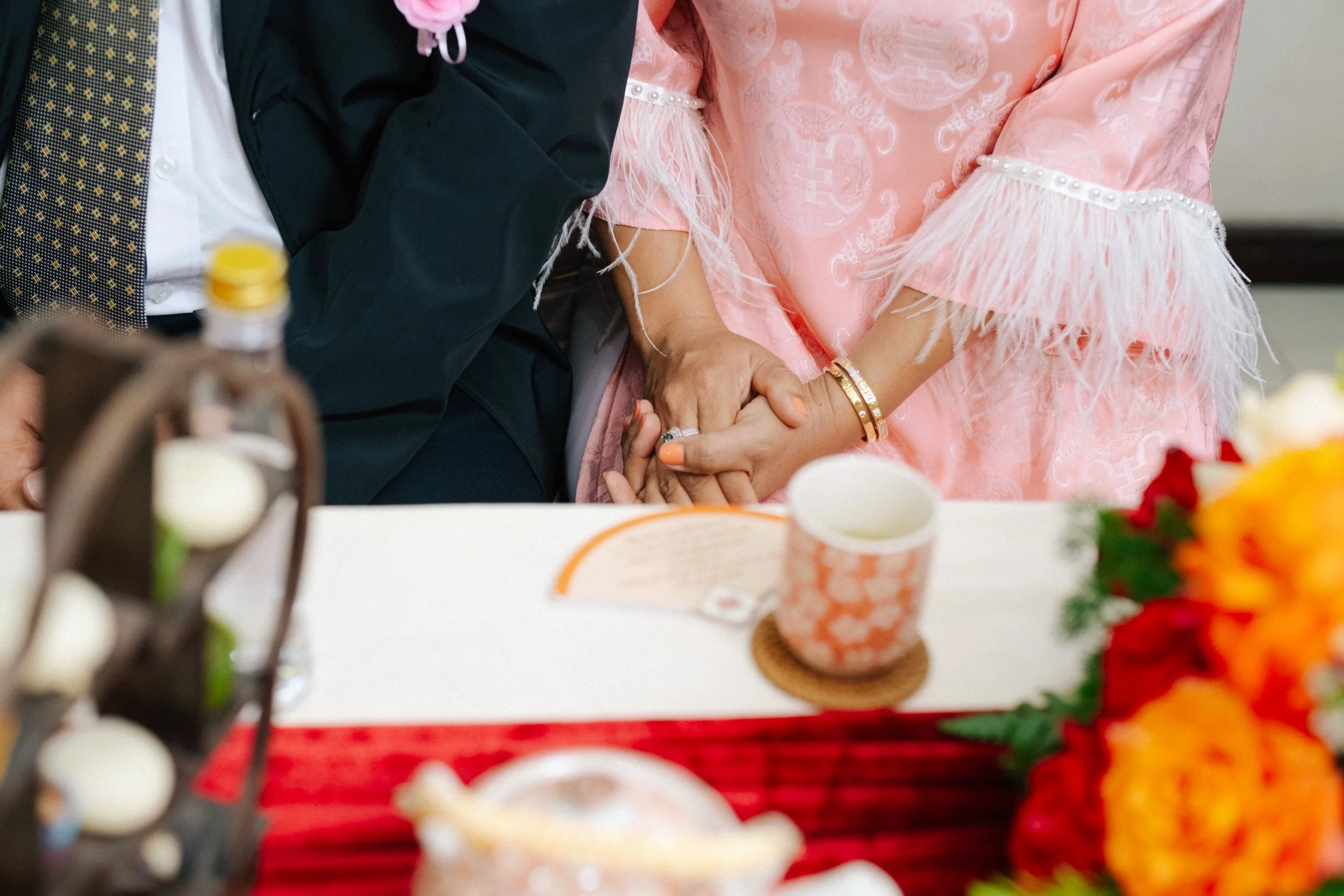 A close-up of two people holding hands at a celebration, with a woman in a pink dress with feathered sleeves and a man in a dark suit. There are decorated tables with flowers and cups in the foreground.