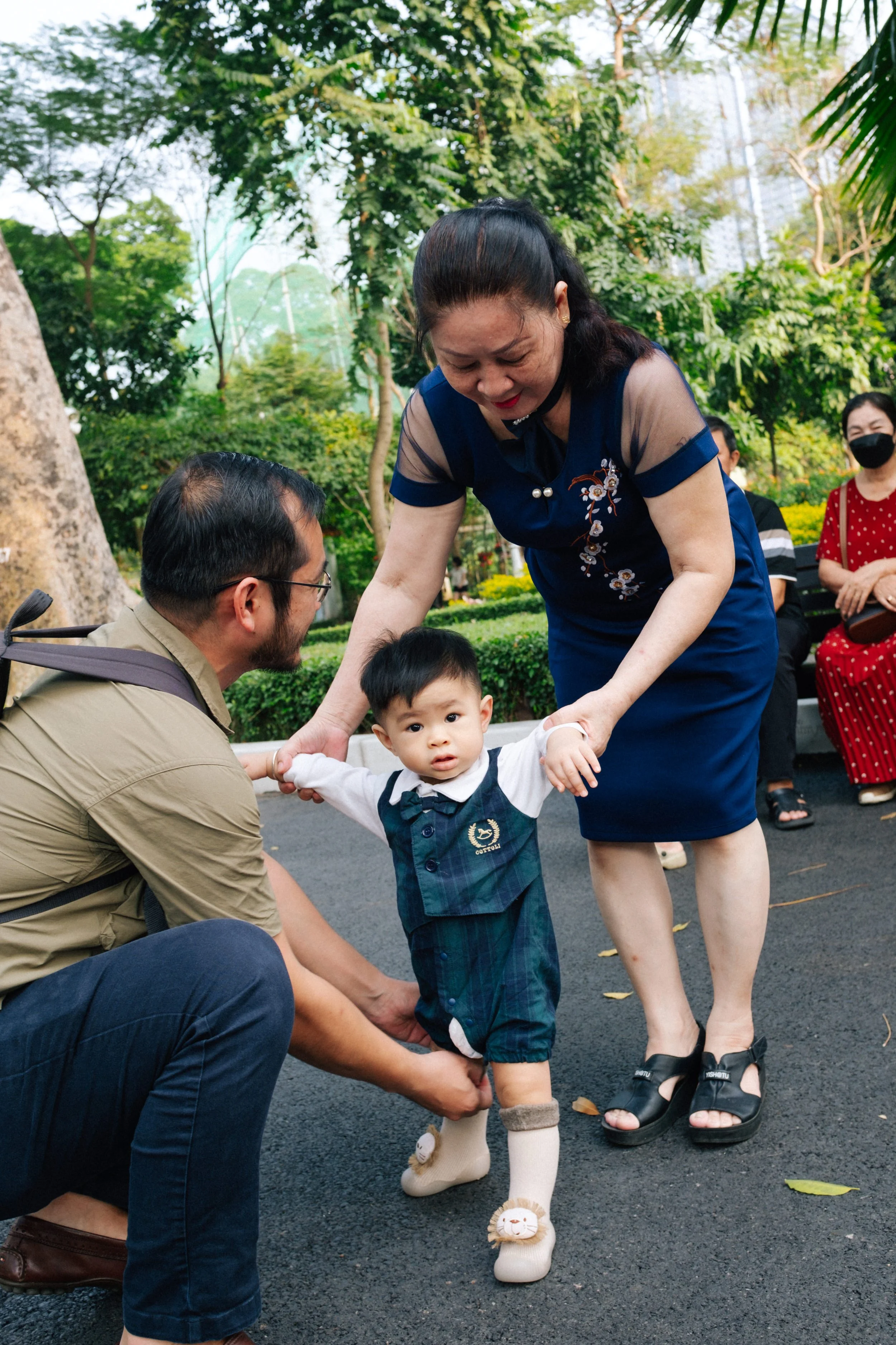 A young boy is being helped to walk by an older woman, with a man assisting him. Several people are sitting on a bench in the background, with trees and greenery surrounding them.