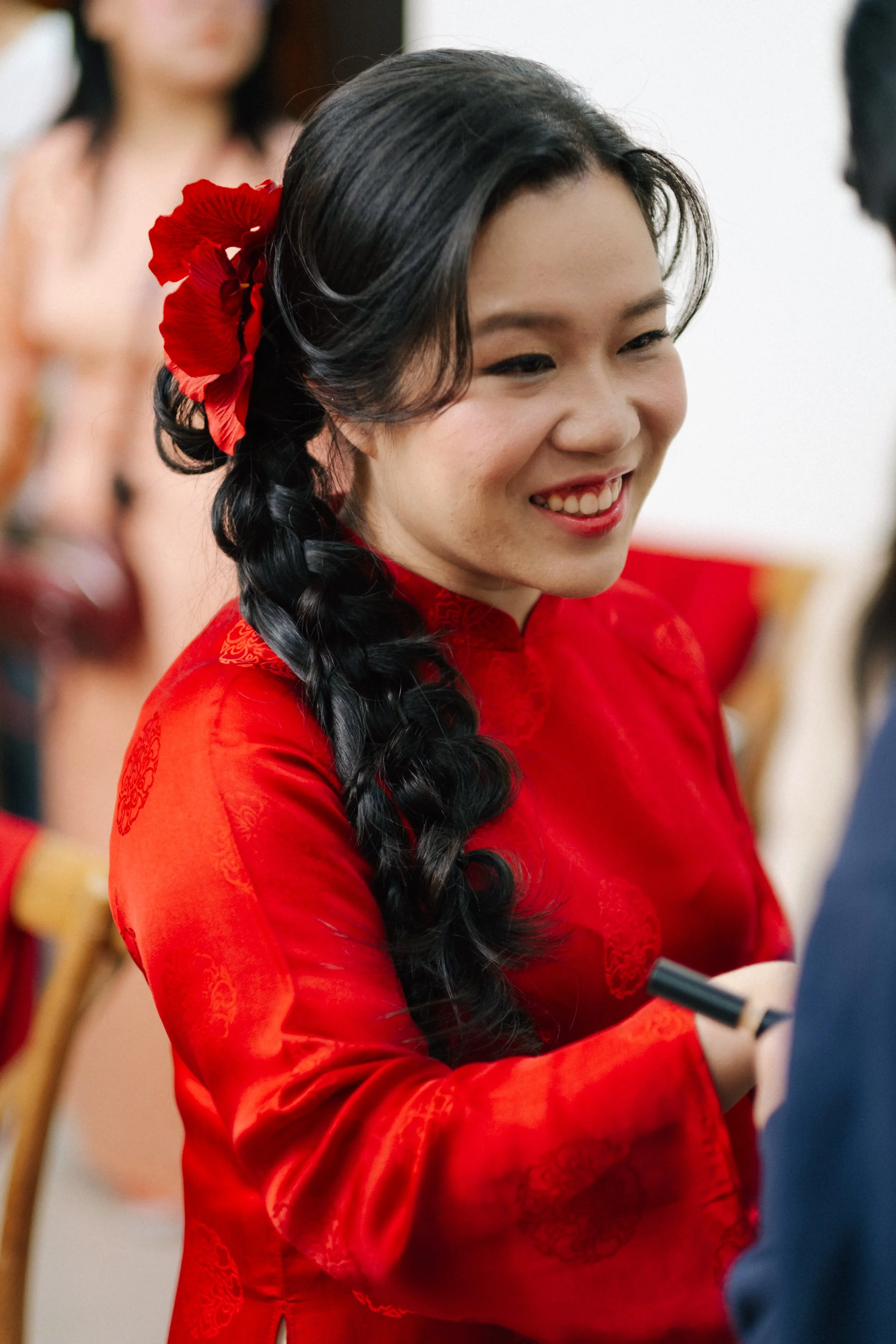 A young woman in a red traditional outfit with a flower in her hair, smiling and engaged in conversation at a cultural event.