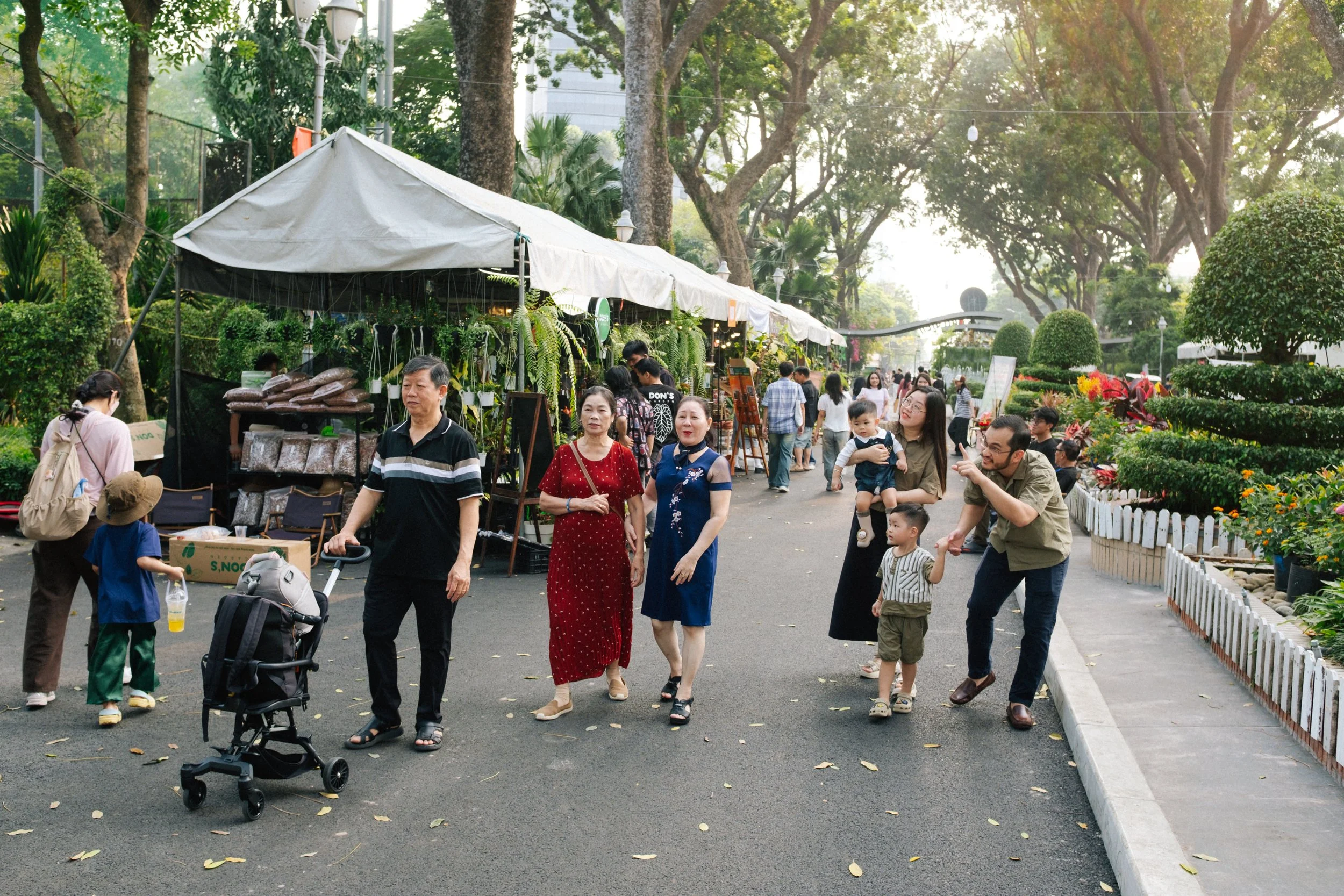 People walking and socializing at an outdoor market surrounded by trees and plants, with vendor stalls selling plants and goods.