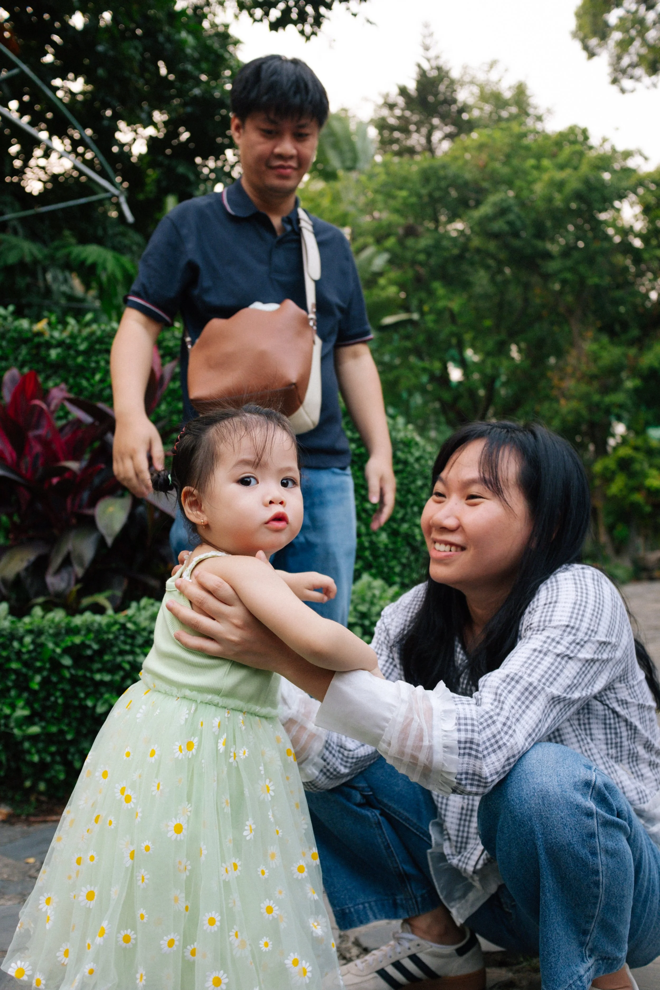 A woman crouching and smiling at a little girl in a yellow floral dress, while a man stands behind them outdoors surrounded by green plants and trees.