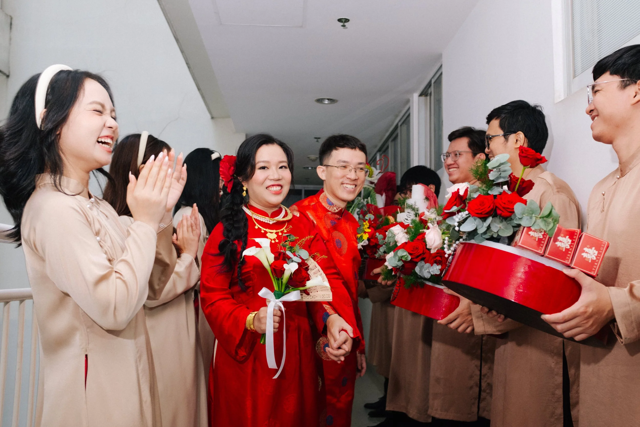 A couple dressed in traditional red Vietnamese wedding attire holding flowers, smiling, surrounded by friends and family in beige outfits, some holding bouquets and gift boxes with red packaging, in an indoor corridor.