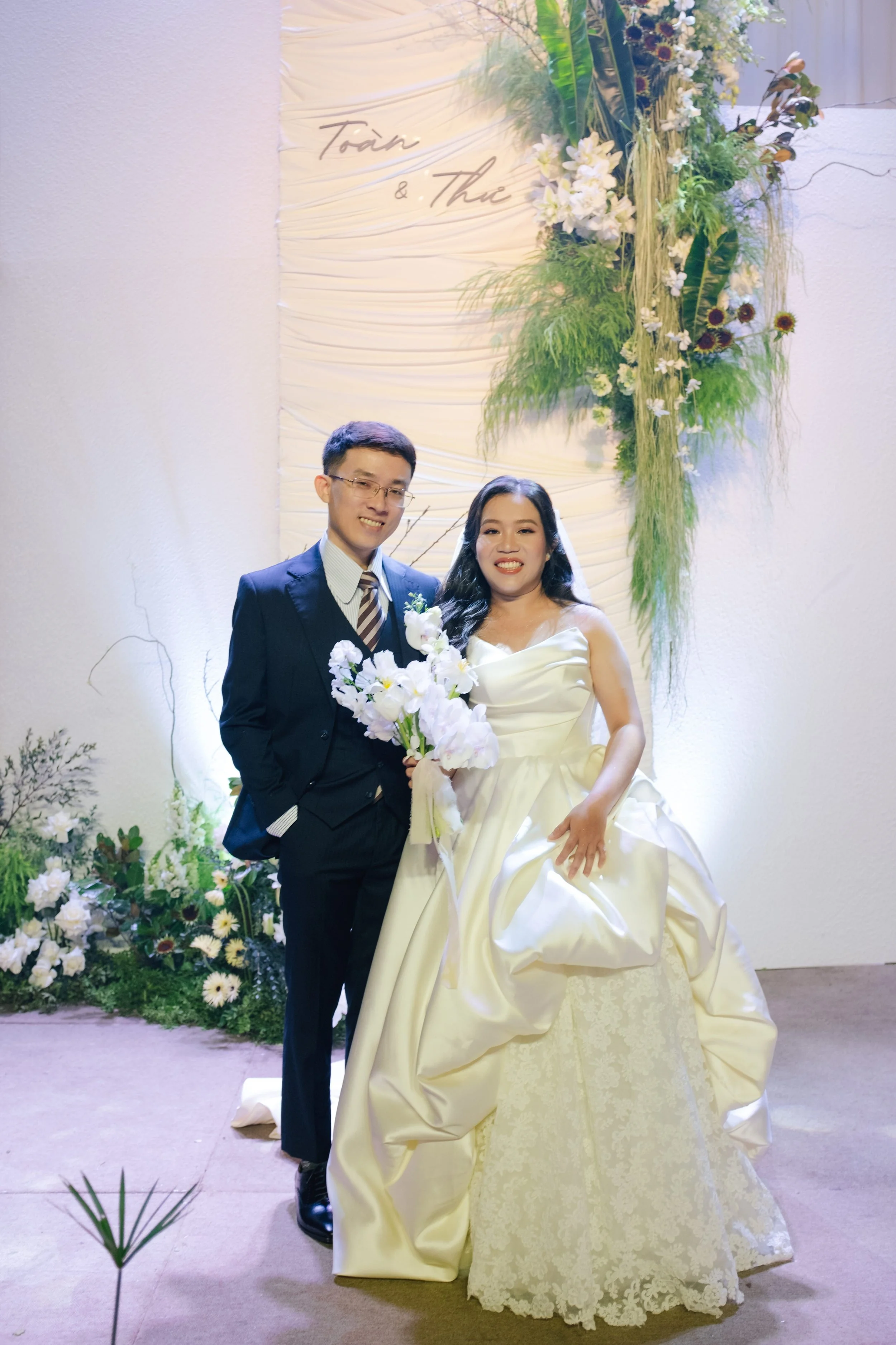 A couple dressed in wedding attire standing in front of a floral backdrop at their wedding venue, with the words 'Tour & The' written on the backdrop.
