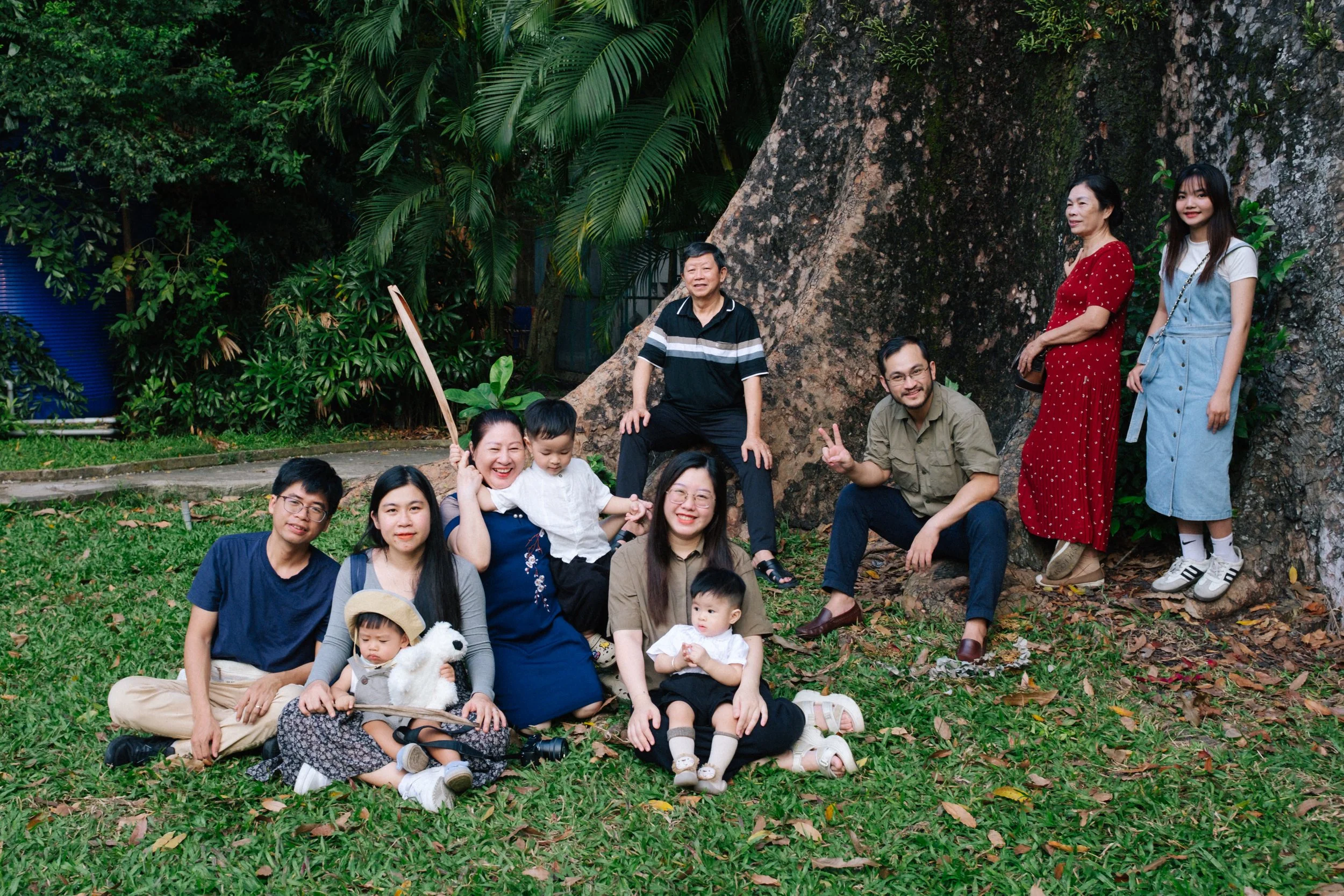 A group of diverse people, including children, adults, and elderly, posing outdoors in front of a large tree. They are smiling, sitting, and standing on grass surrounded by lush greenery.
