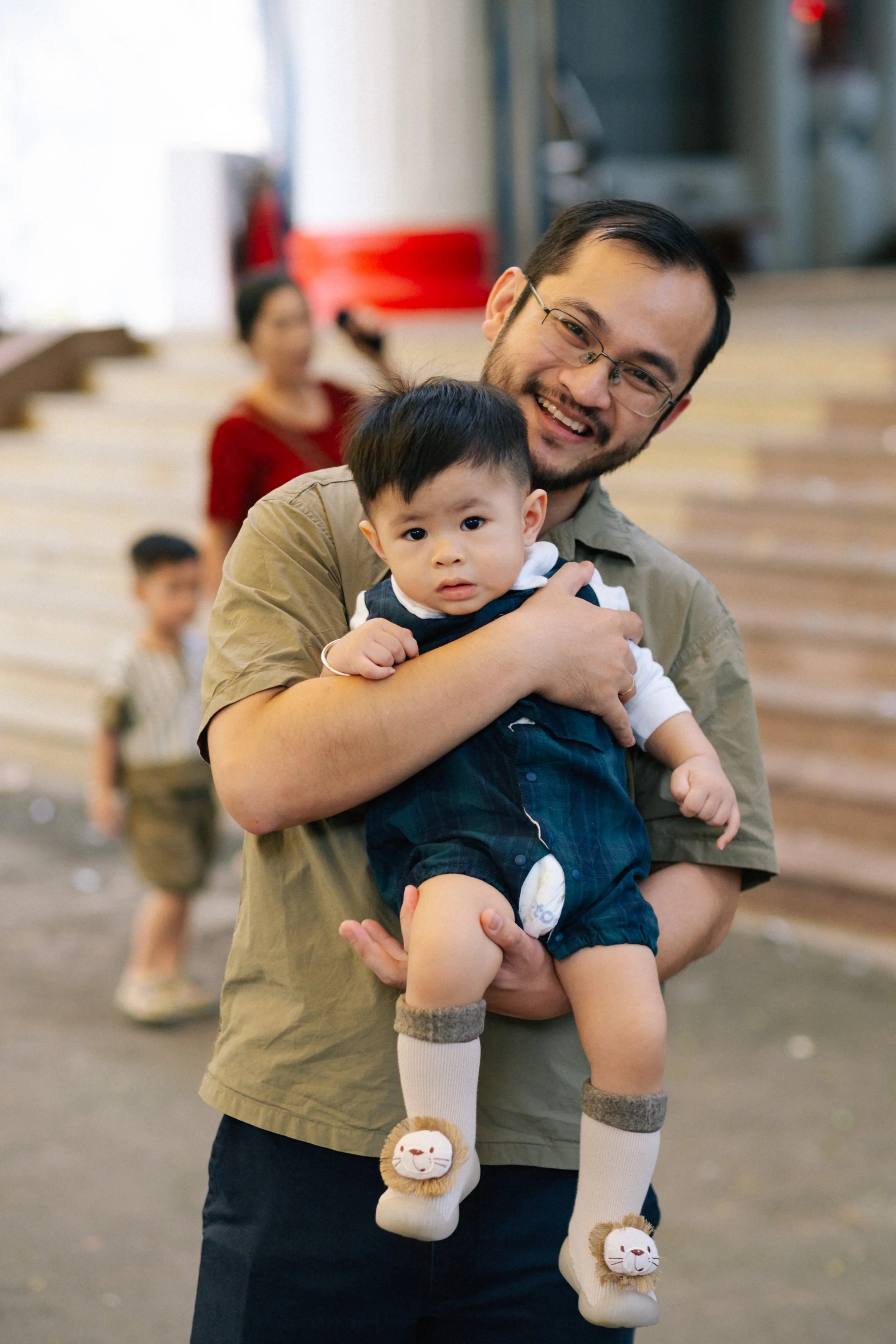 A smiling man holding a young child in his arms, with a blurred background of two children and an adult in an indoor setting.