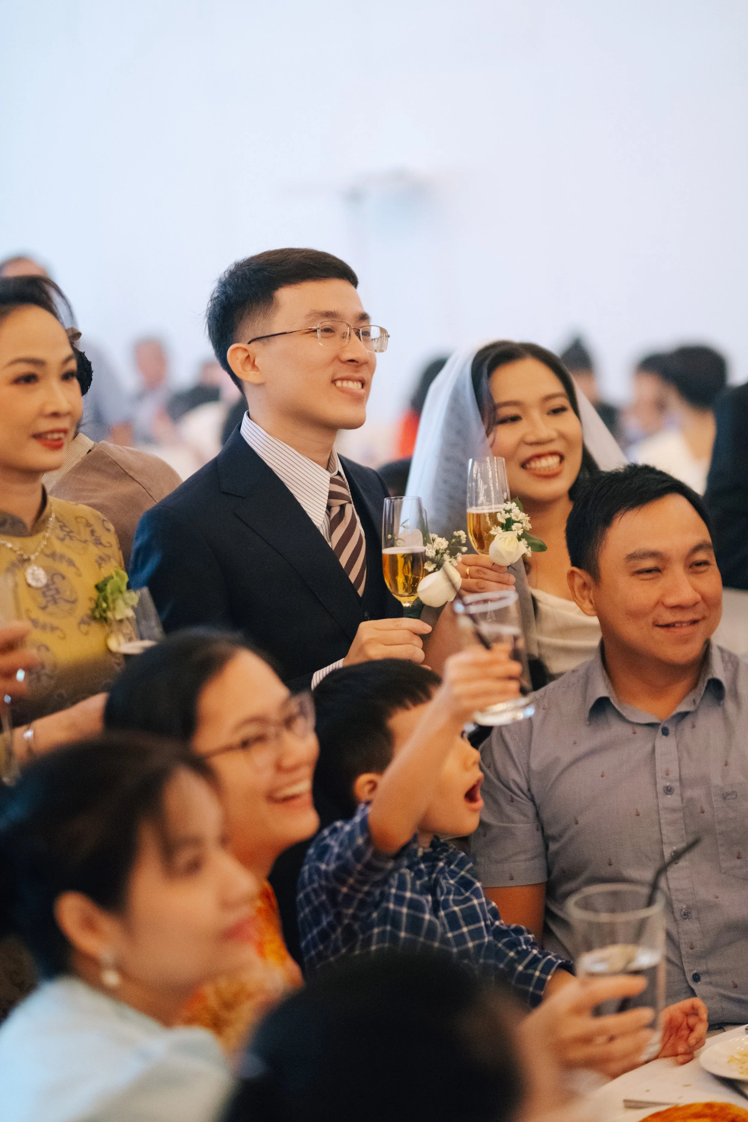 Group of people celebrating at a wedding reception, including a man and woman with a veil, holding glasses of champagne.