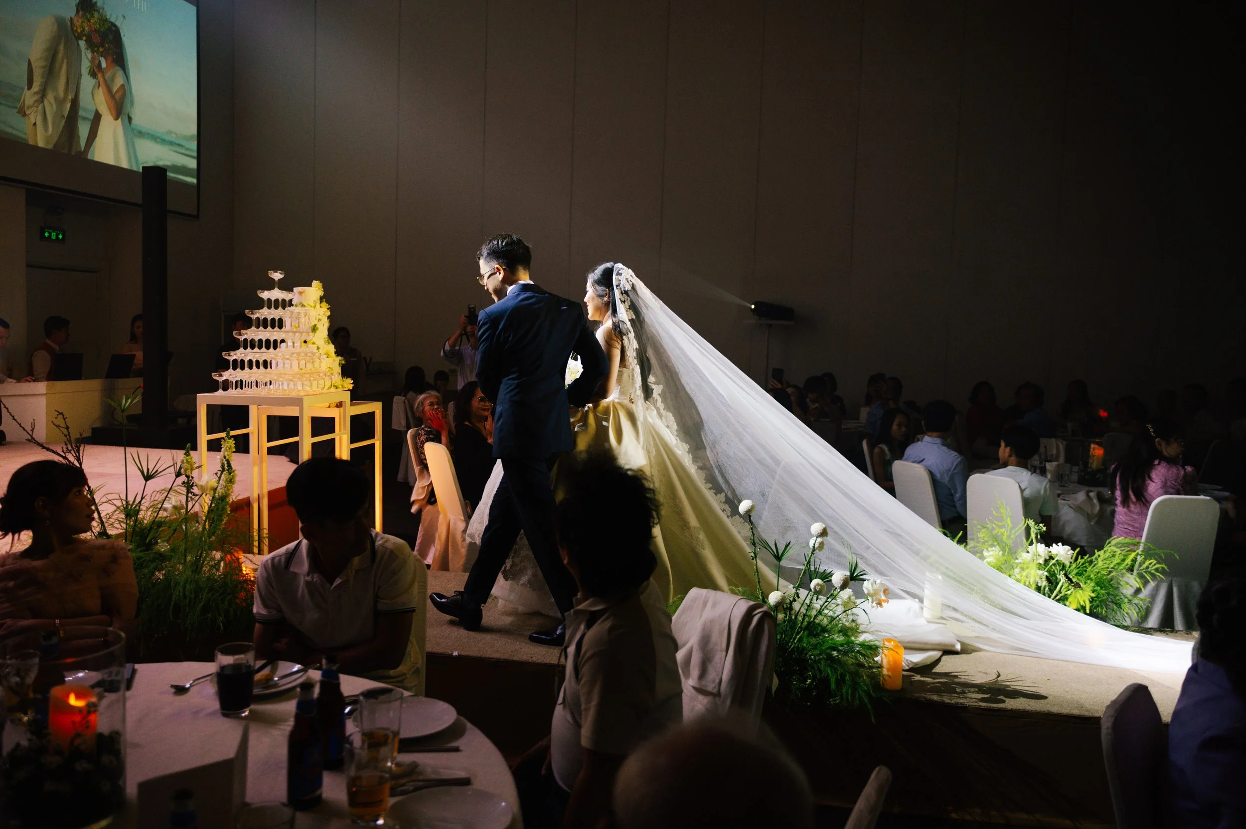 A bride and groom walking across a stage at a wedding reception, surrounded by seated guests and floral decorations, with a multi-tiered wedding cake in the background.