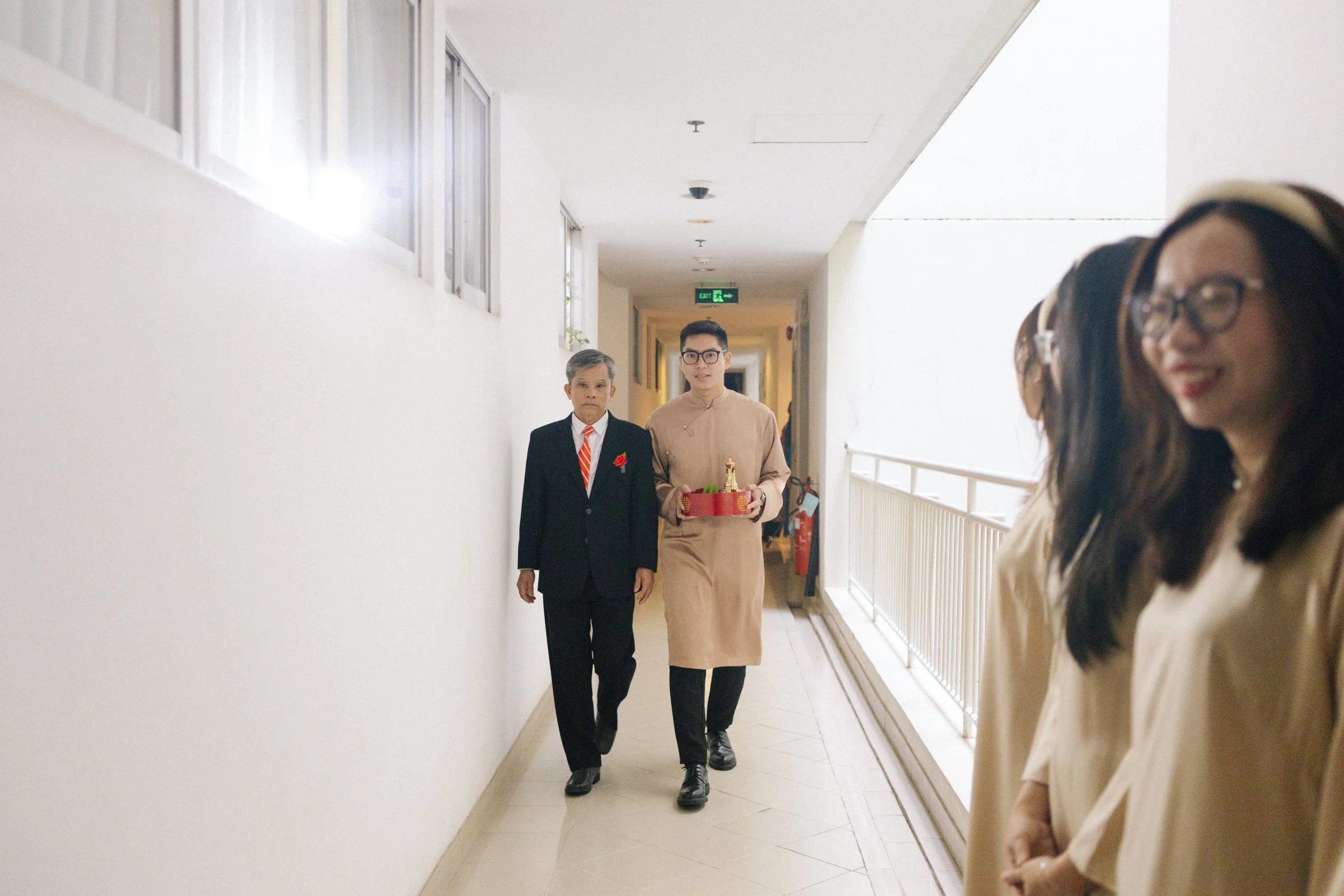 A young man in traditional attire carrying a red tray with a small Buddha statue, walking in a corridor alongside an older man in a suit, with three women wearing beige dresses and glasses standing on the right side.