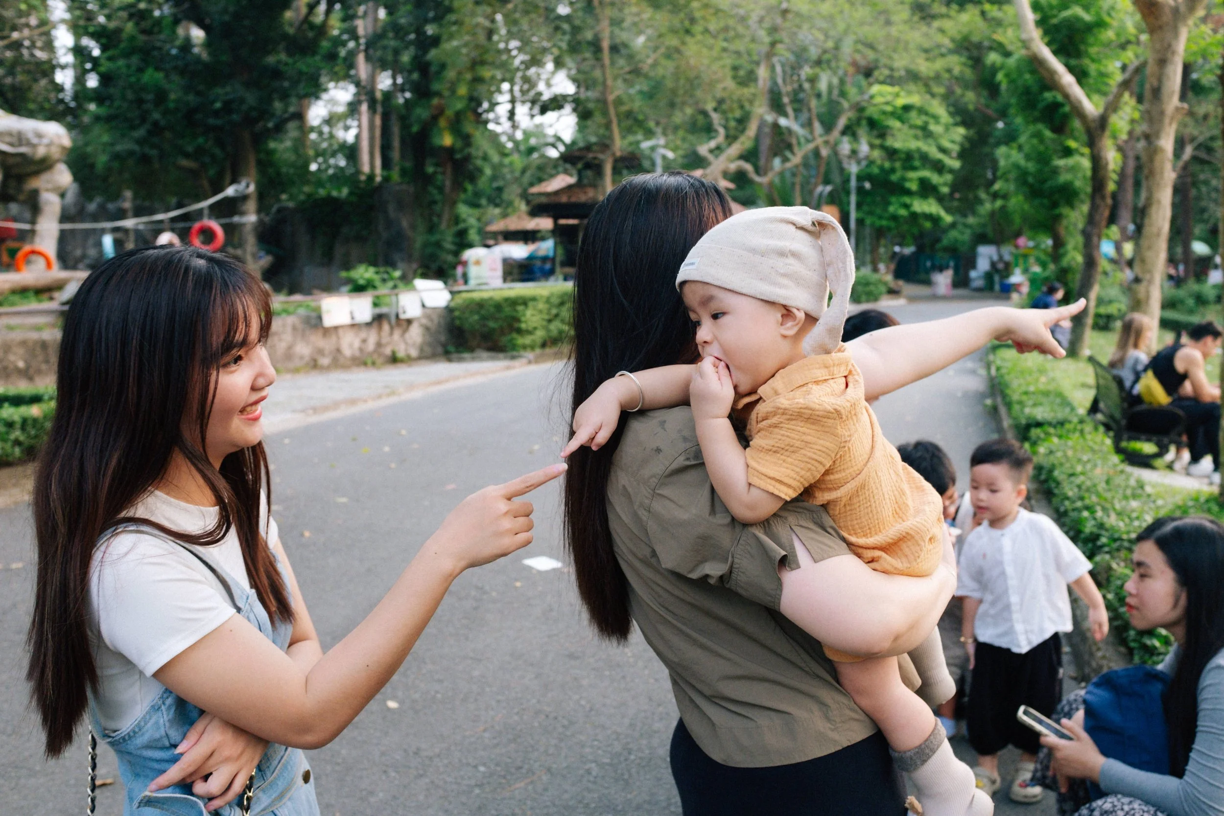 A woman holding a young child in a park, with another girl pointing at the child. Several other children and adults are in the background, and trees surround the scene.